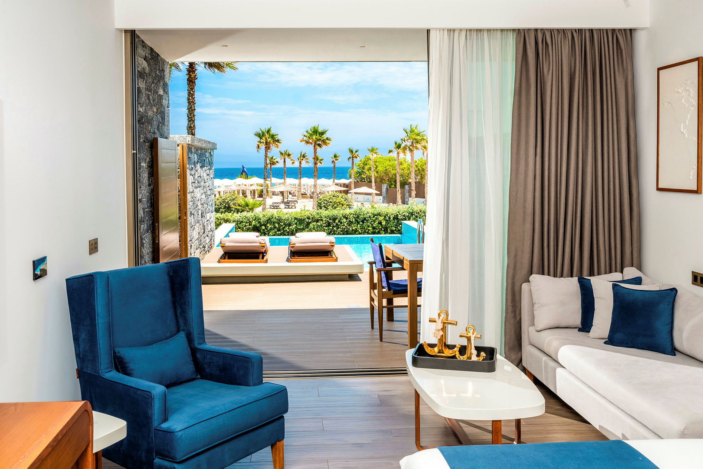 Vy från ett hotellrum som vetter mot en terrass med två solsängar, en pool och en havsutsikt med palmer. (AI-genererad text)
