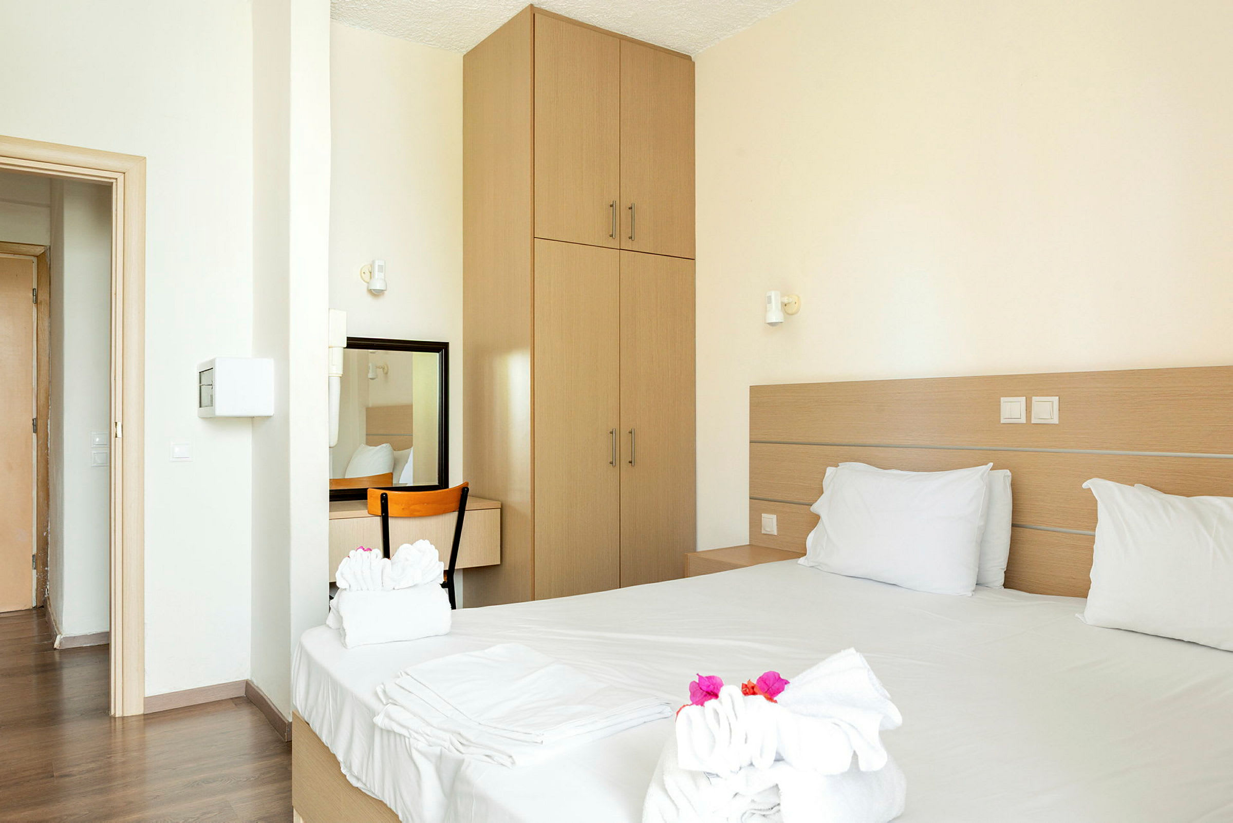 Ett hotellrum med en dubbelsäng, vit sängkläder och handdukar dekorerade med blommor, en garderob, en stol och ett skrivbord med spegel. (AI-genererad text)