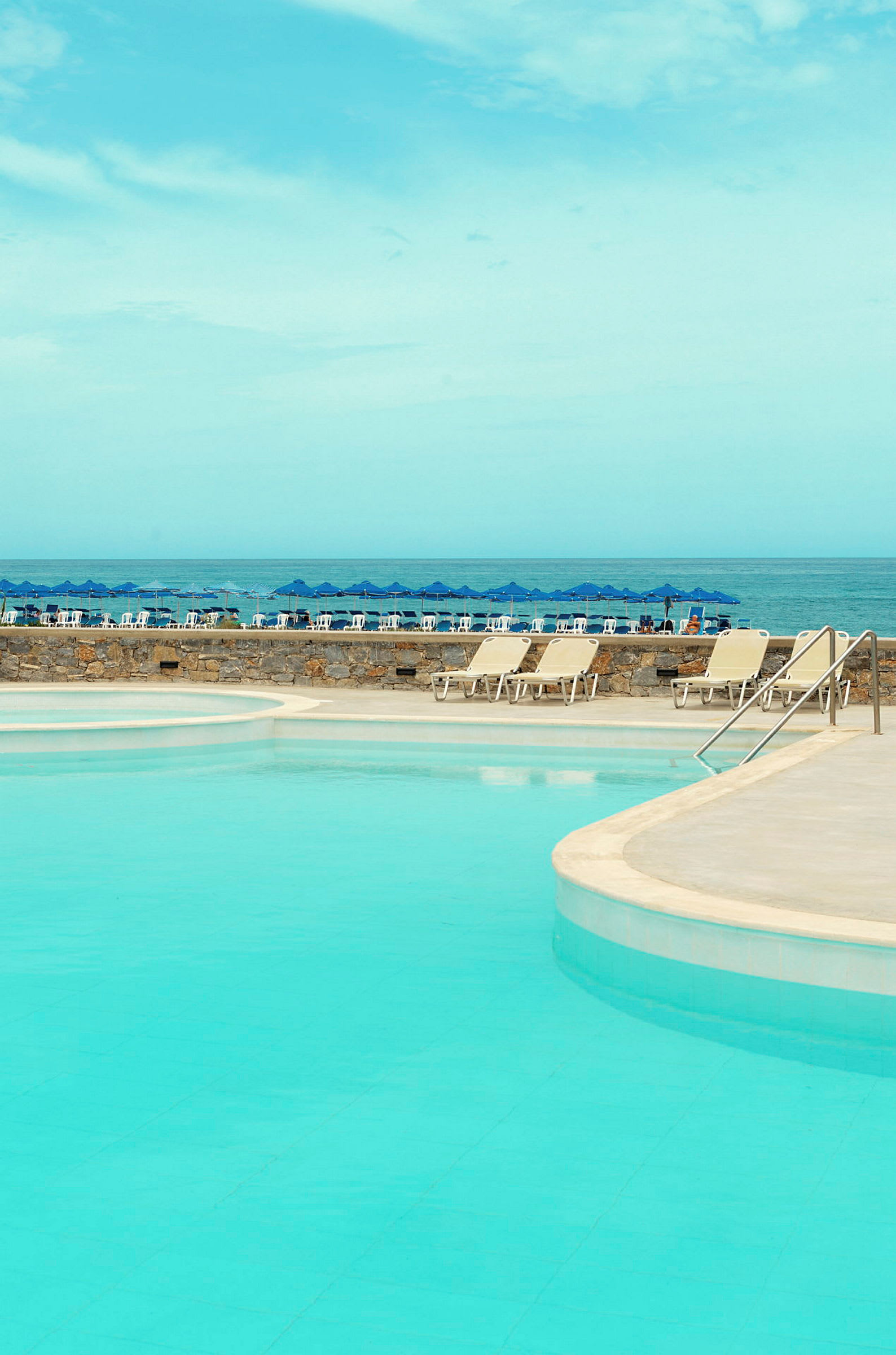 En pool med klarblått vatten och solsängar vid kanten, samt en blå himmel och blå parasoller vid havet i bakgrunden. (AI-genererad text)
