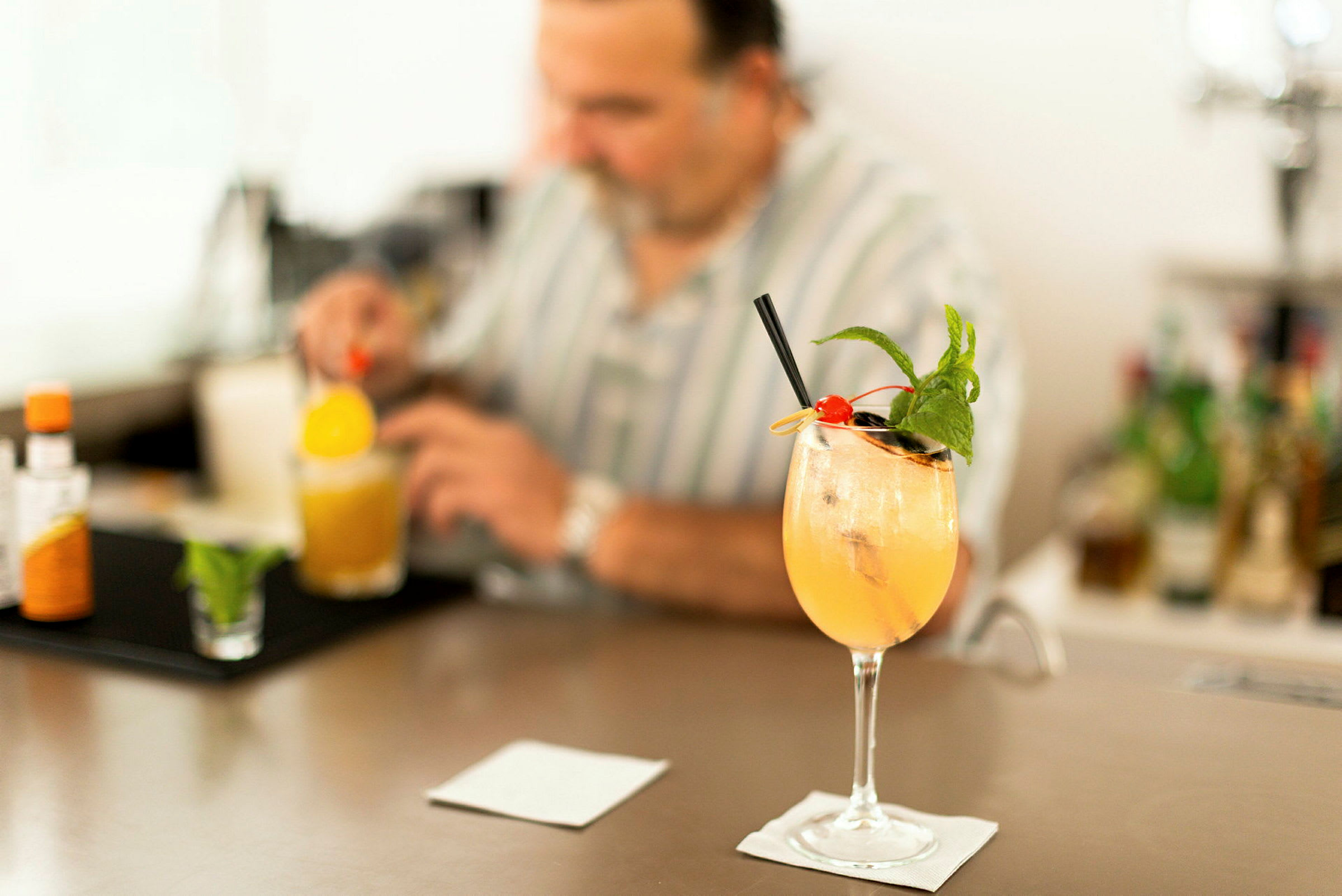 En cocktailglas med en färgglad dryck, toppad med is, mynta och en körsbär, står på en bardisk. En person i bakgrunden blandar en drink. (AI-genererad text)
