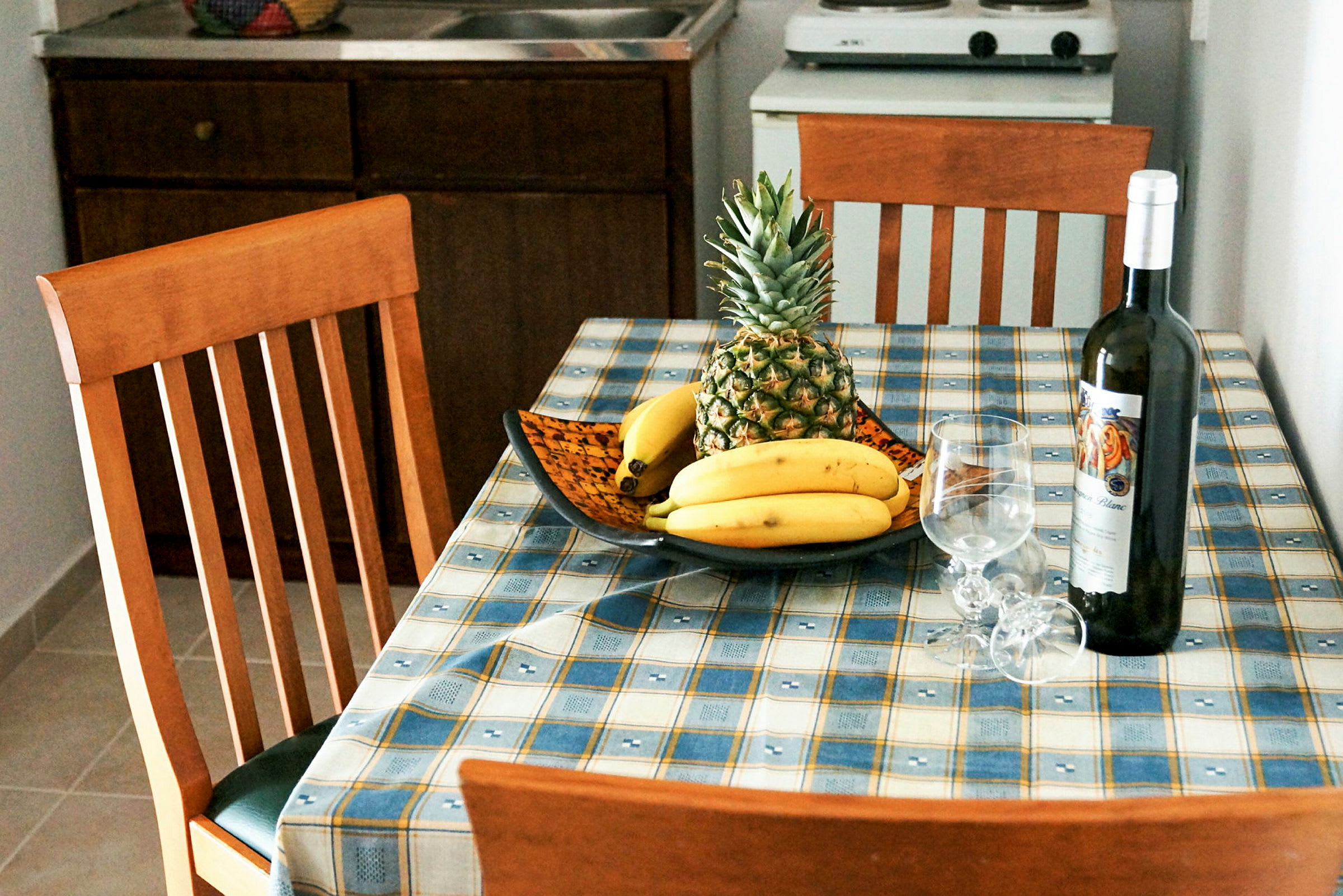 Ett köksbord med en fruktskål med ananas och bananer, samt en vinflaska och två vinglas. (AI-genererad text)