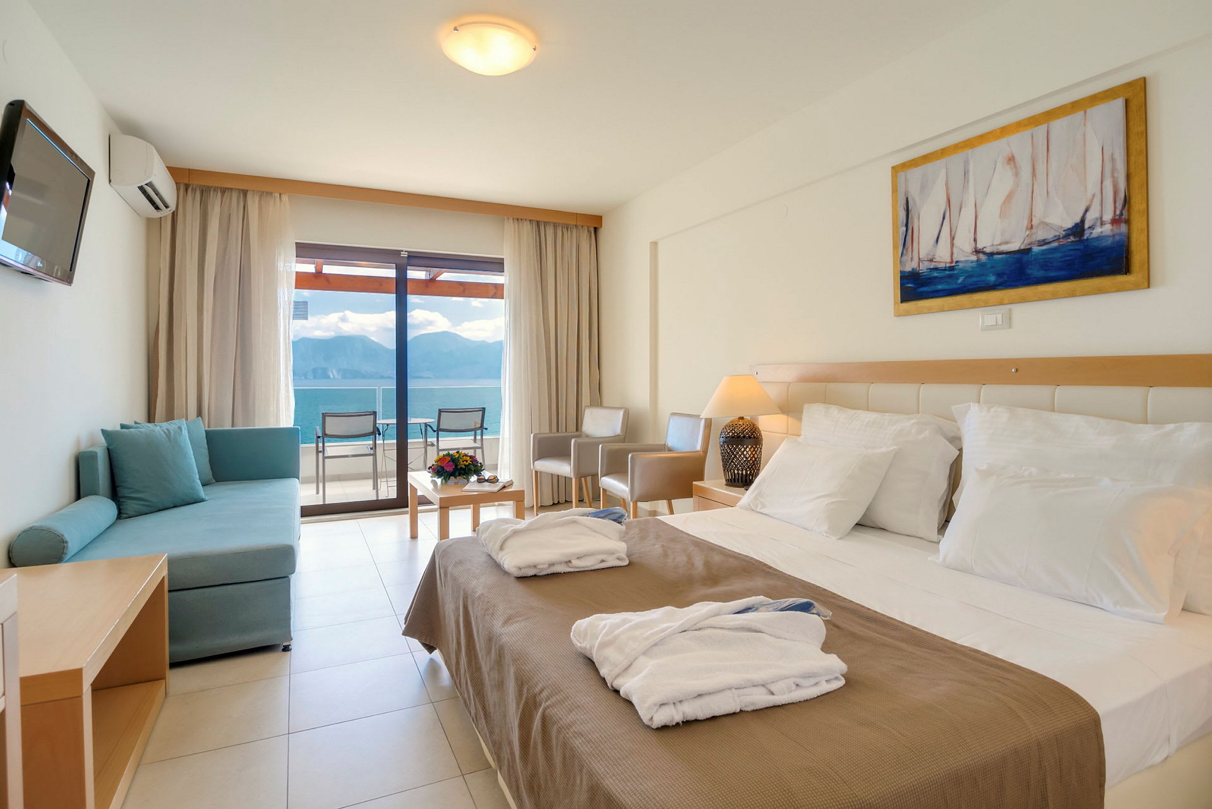 Ett hotellrum med en dubbelsäng, en soffa, ett bord med blommor och en utsikt mot havet genom glasdörrar. (AI-genererad text)