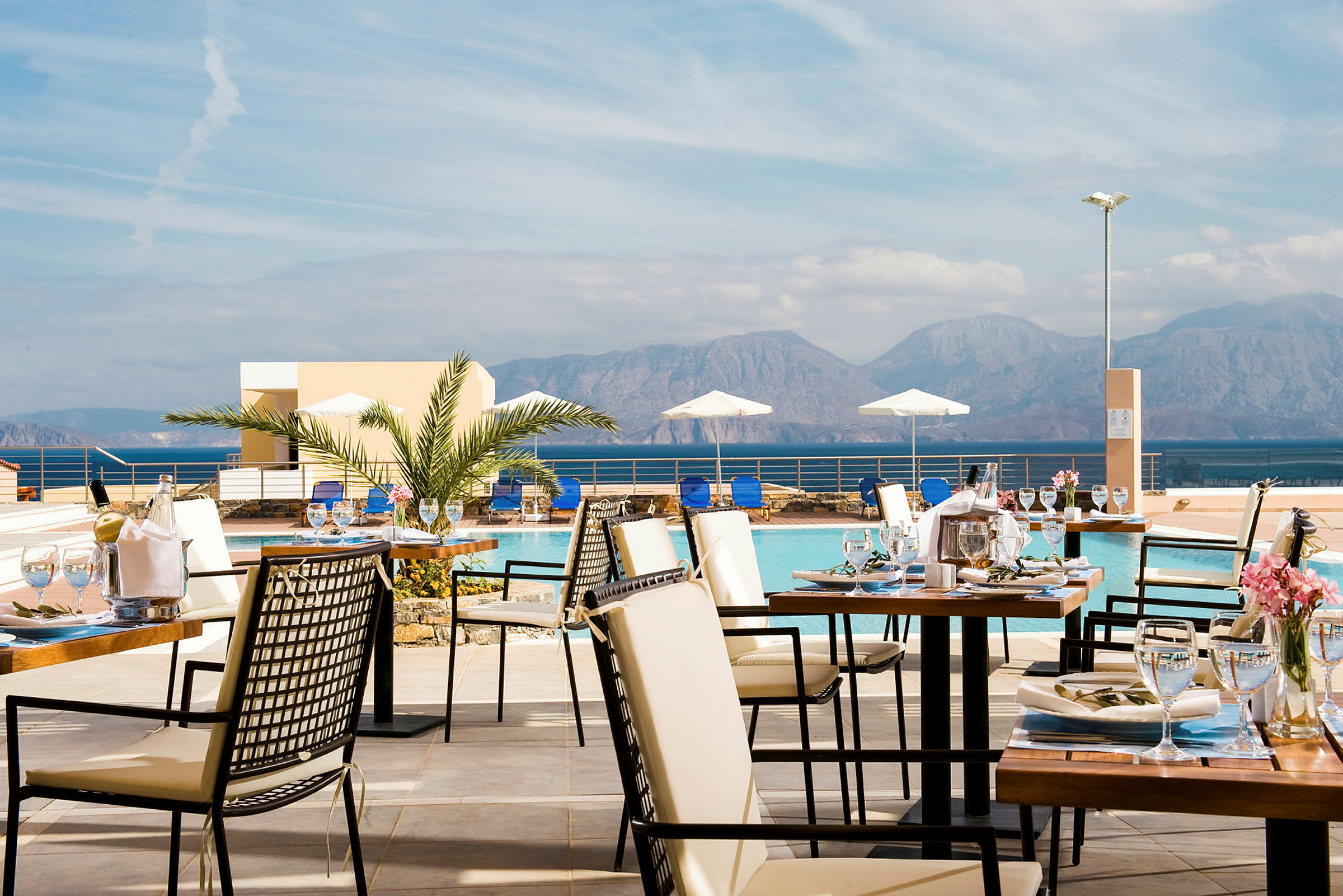 I förgrunden finns uppdukade matbord utomhus med glas och servetter. I bakgrunden syns en pool, solstolar och parasoller samt berg i fjärran. (AI-genererad text)