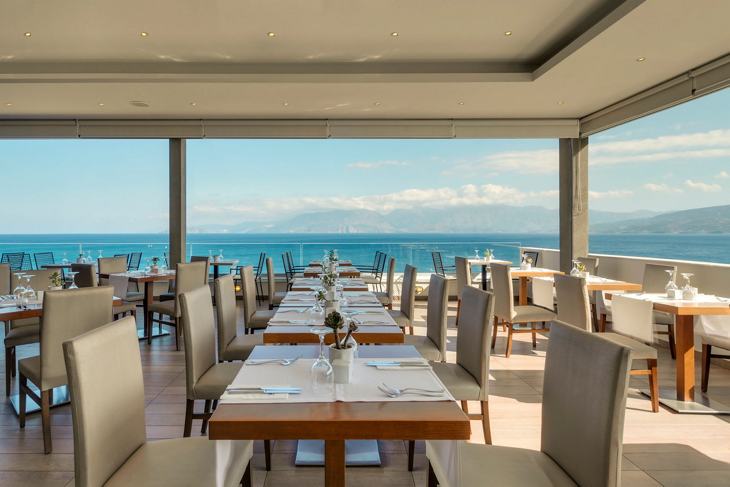 Utsikt över en restaurang med dukade bord och stolar, med havet och berg i bakgrunden. (AI-genererad text)
