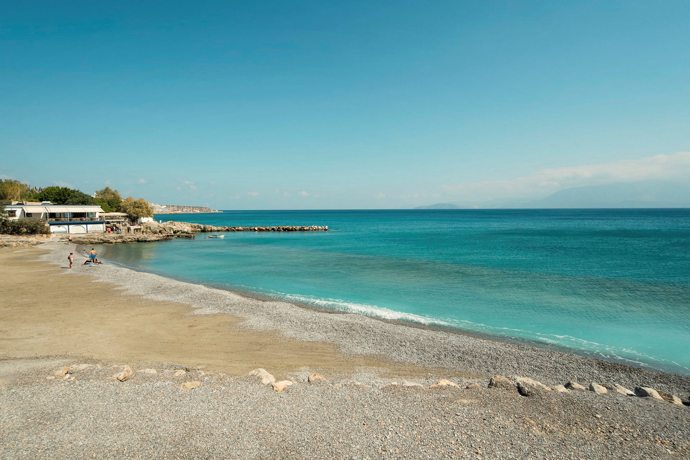 Strand med småsten och klarblått vatten, med några människor nära en byggnad. (AI-genererad text)