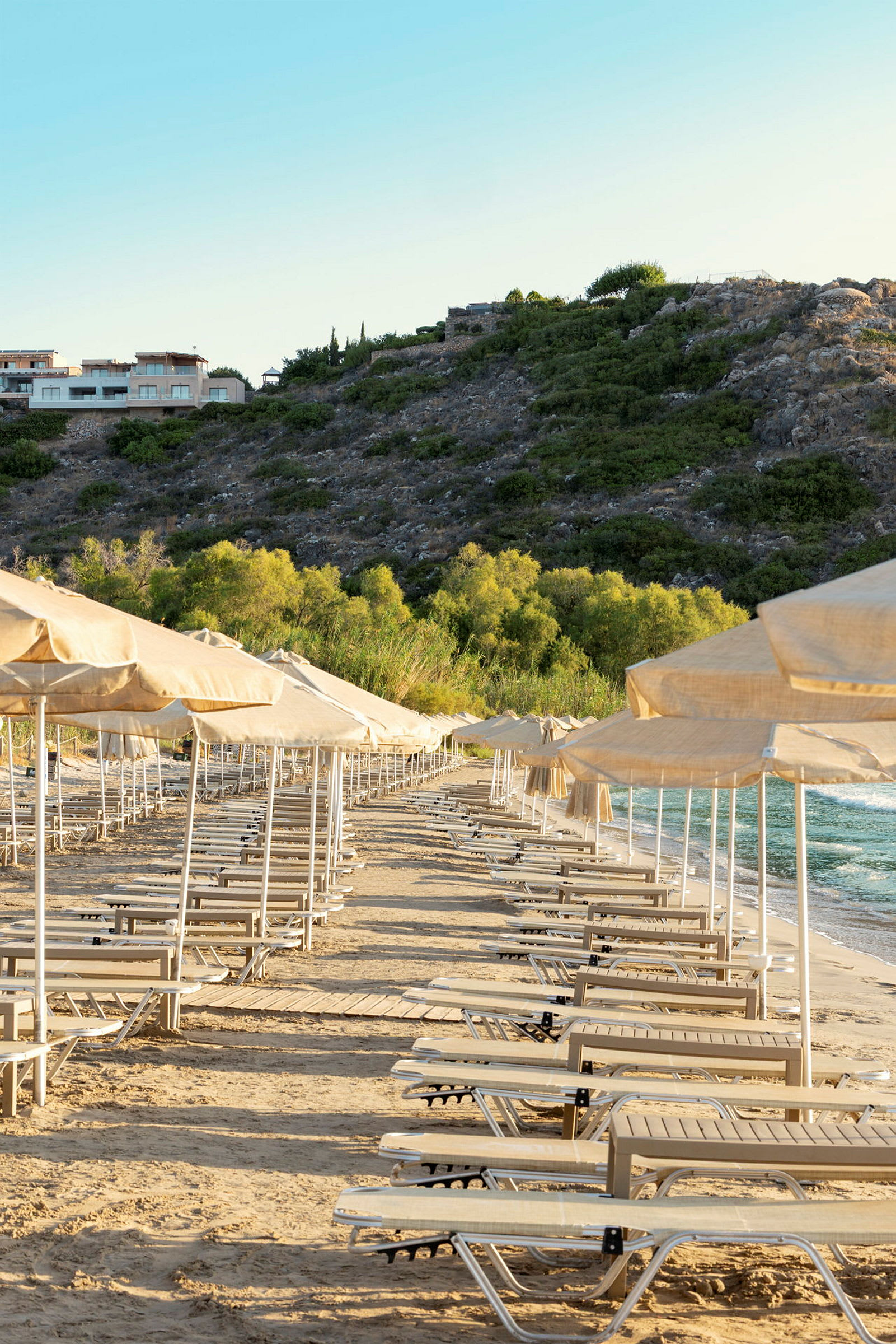 En strand med solstolar och parasoller uppställda i rader, omgivna av grönska och berg. (AI-genererad text)