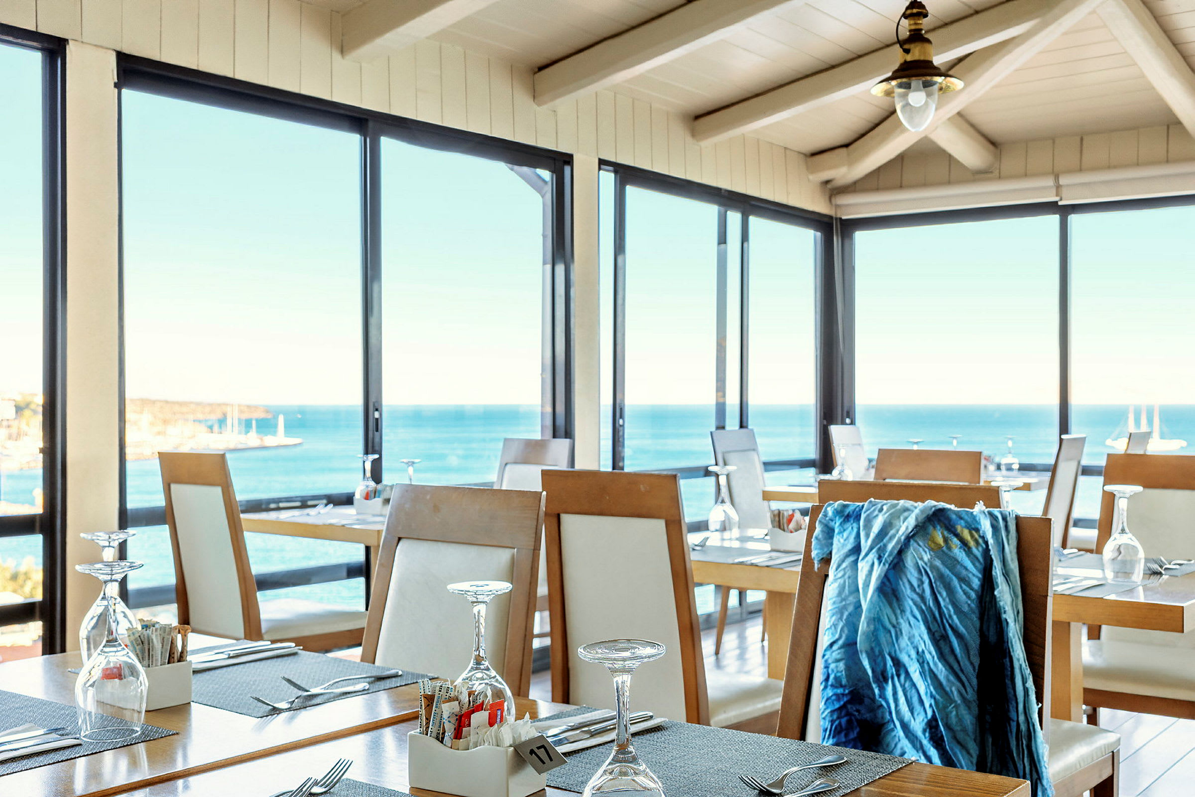 Ett ljust och luftigt restaurangområde med stora glasfönster som ger utsikt över havet. Bord är dukade med glas och bestick. (AI-genererad text)