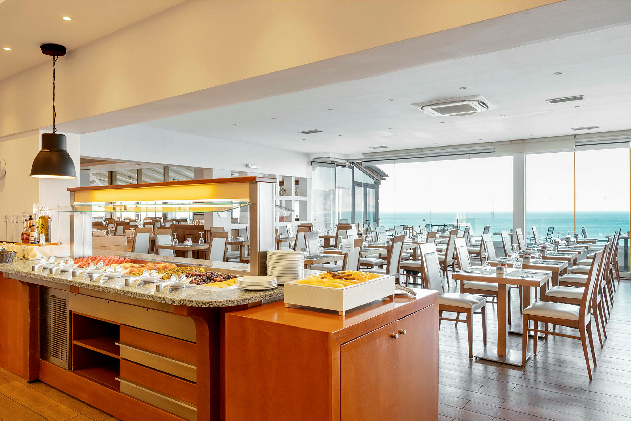 Bild av en restaurang med buffébord och ljusa stolar. Havet syns genom fönstren. (AI-genererad text)