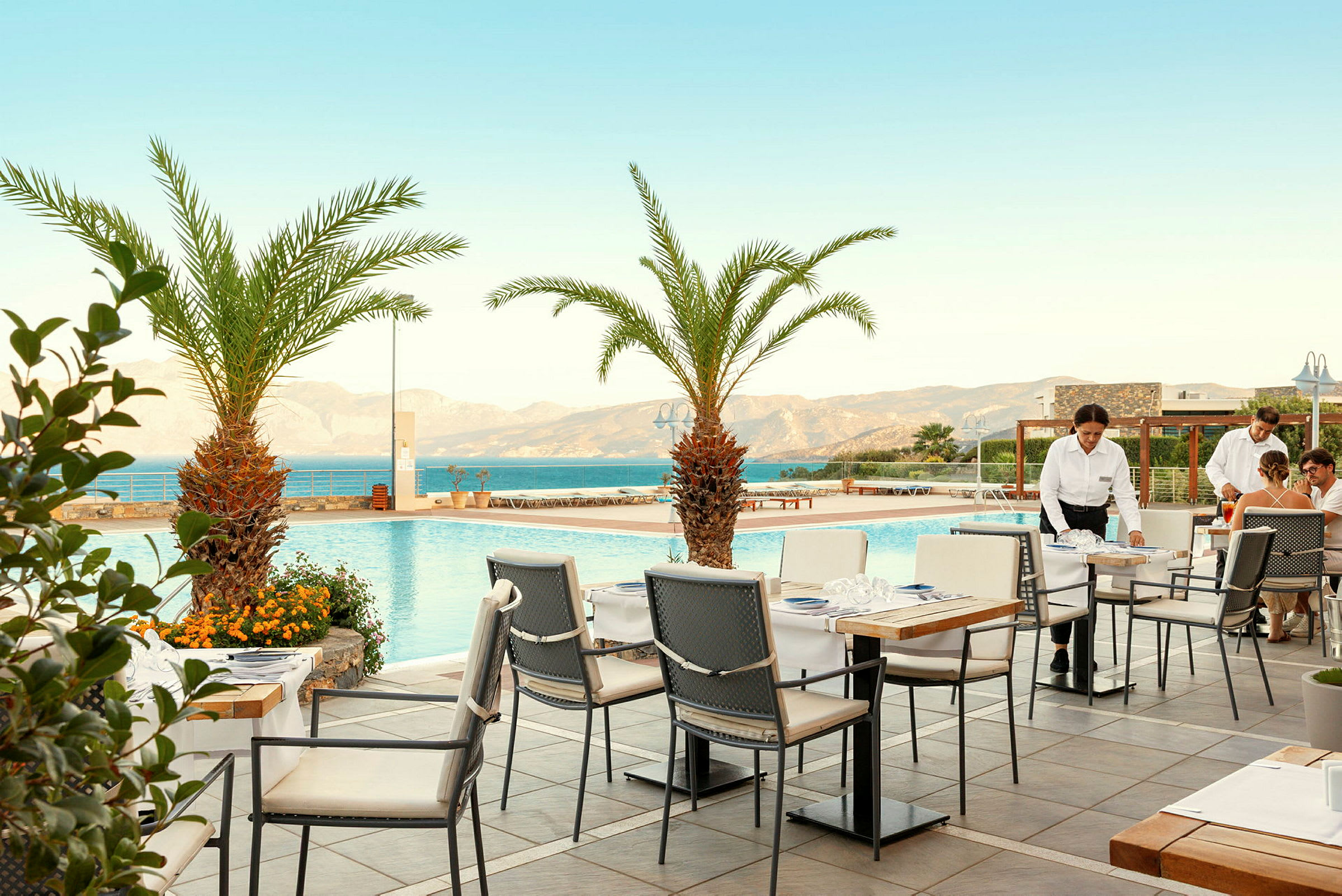 En terrass med utsikt över en pool och havet, omgiven av palmer. Bord och stolar är placerade på terrassen, och personal serverar gäster. (AI-genererad text)