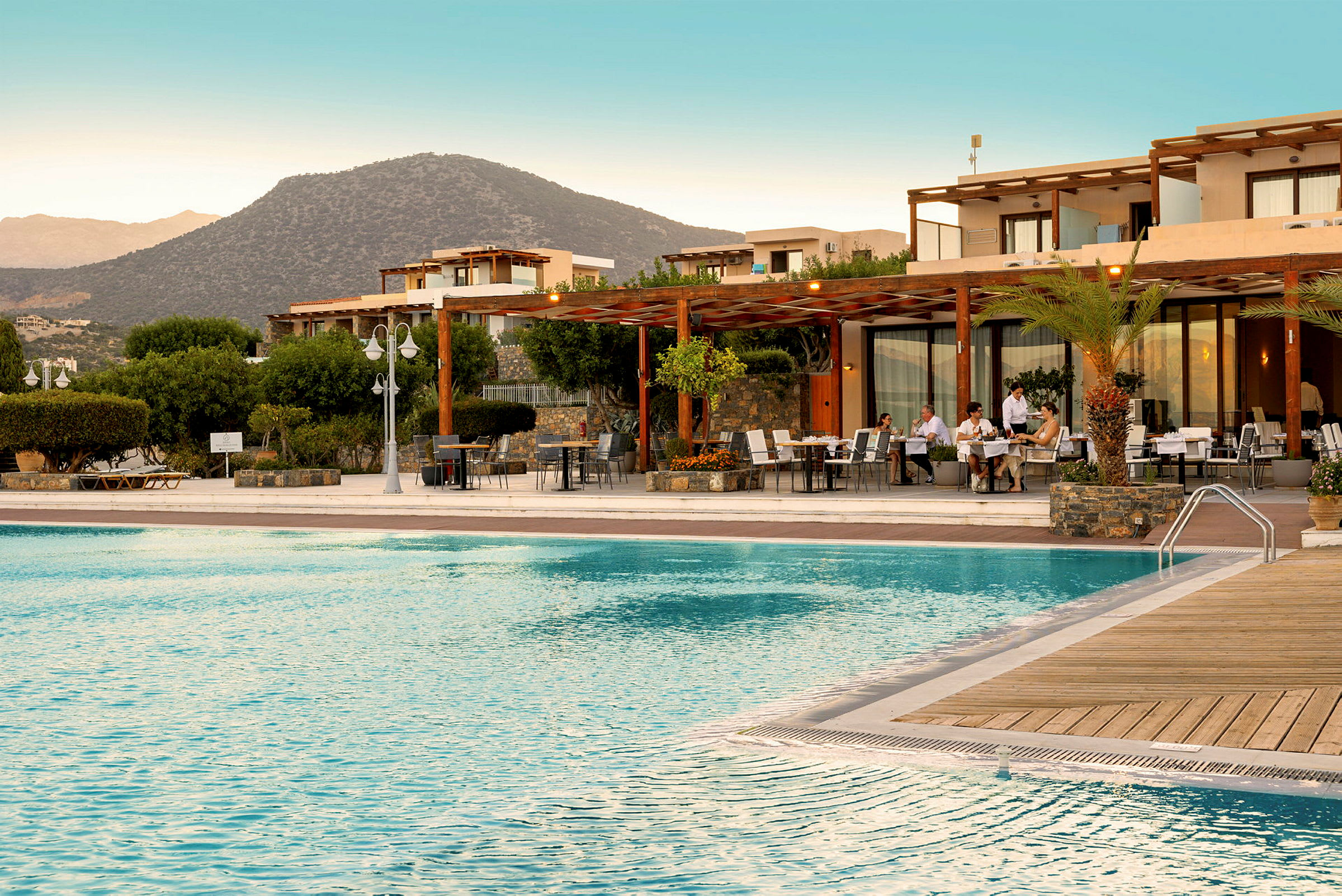En hotellpool med solstolar och en terrass där gäster sitter vid bord. I bakgrunden syns byggnader och berg. (AI-genererad text)