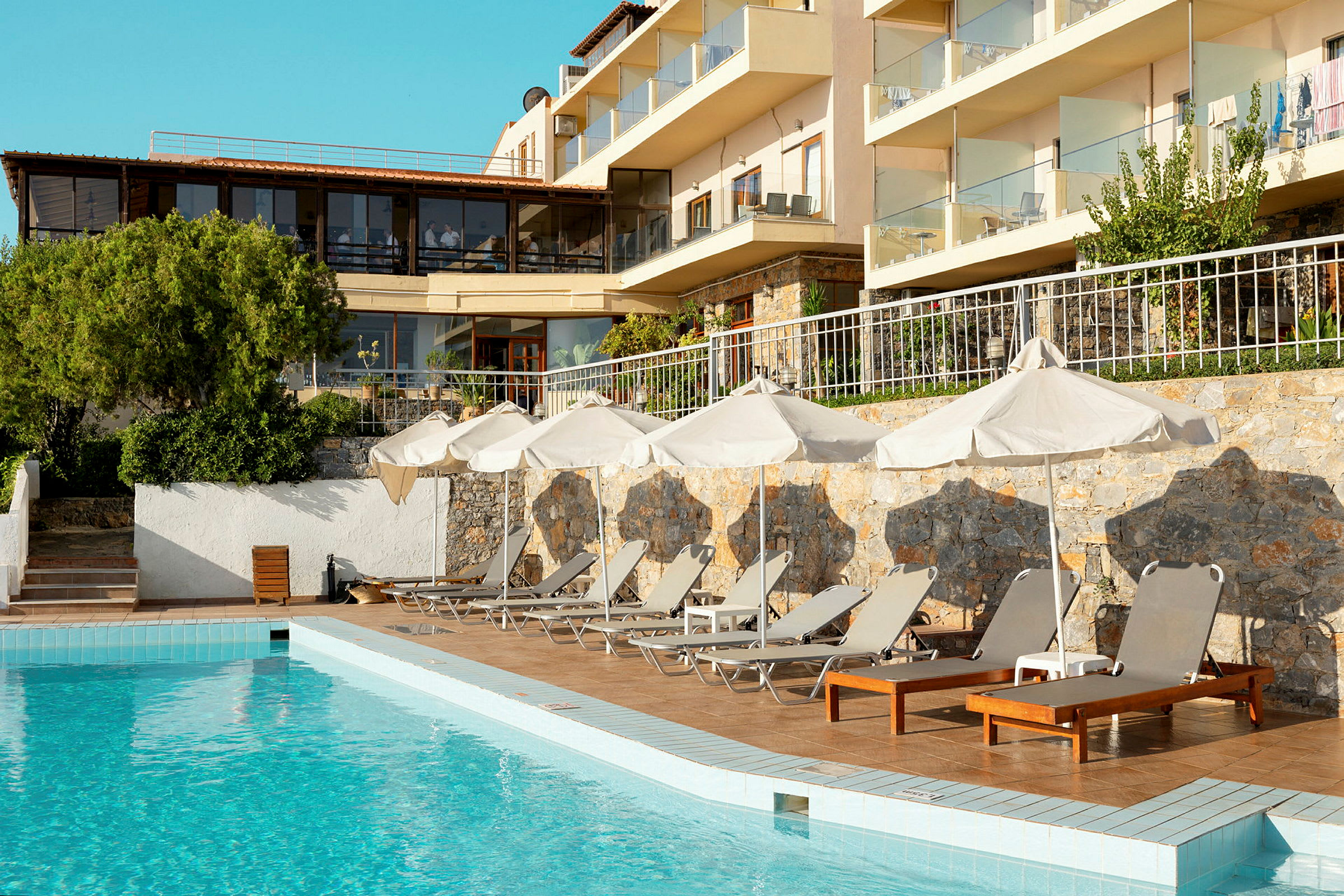 En hotellpool omgiven av solstolar och parasoller med en byggnad i bakgrunden. (AI-genererad text)