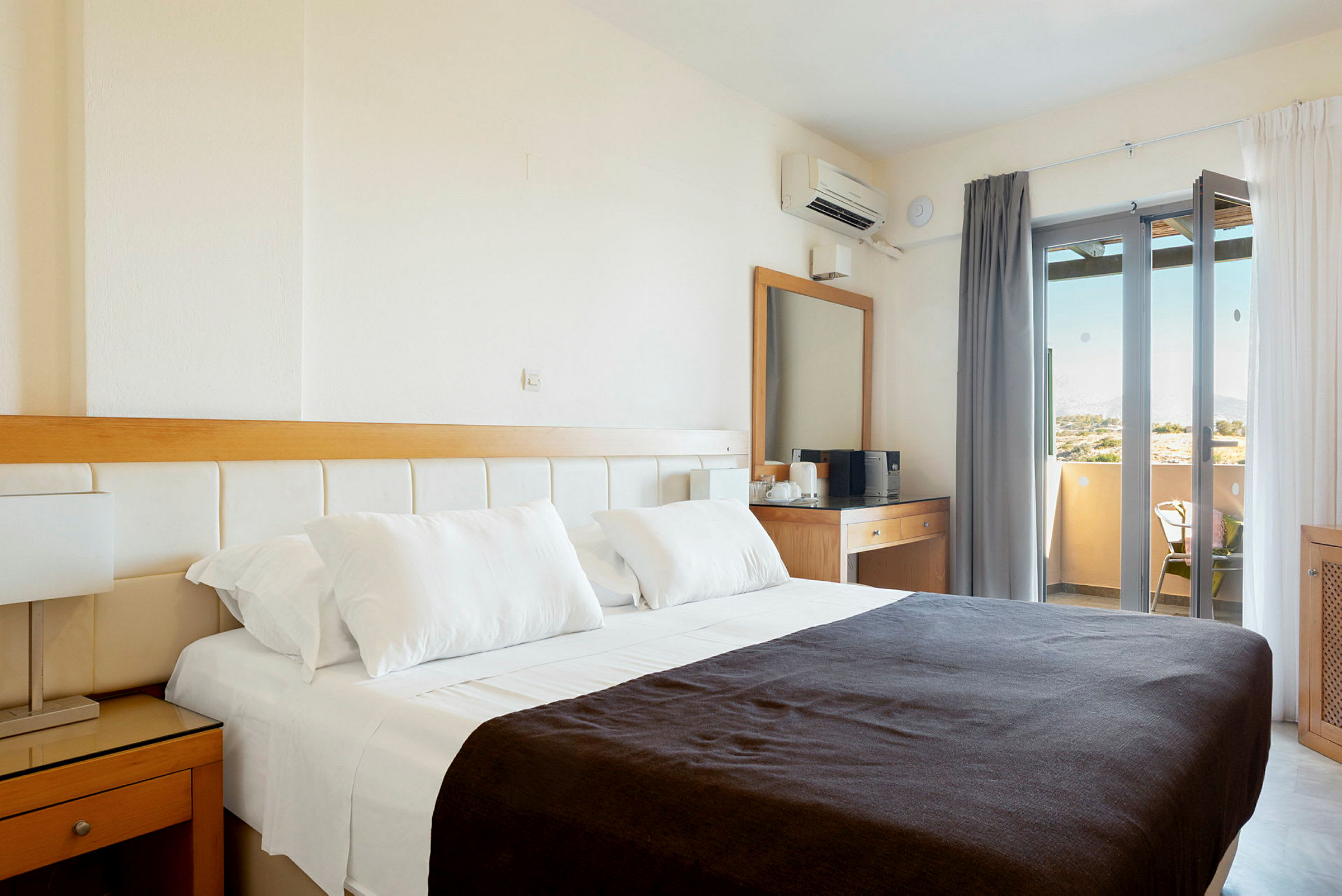 Ett hotellrum med en dubbelsäng klädd i vita lakan och ett mörkt täcke. I rummet finns också ett skrivbord med spegel och en balkong med utsikt. (AI-genererad text)