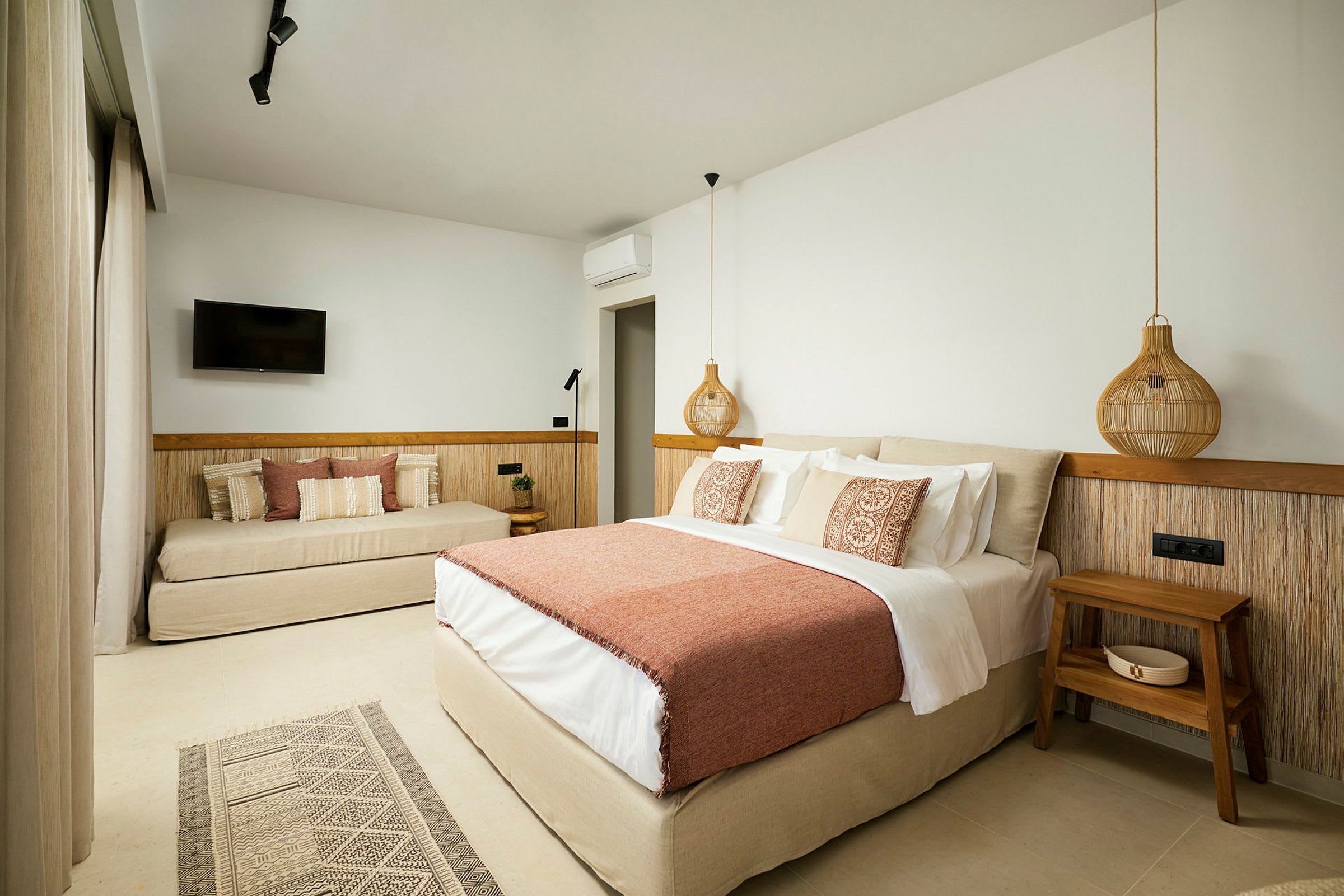 Ett hotellrum med en stor säng med orange överkast och dekorativa kuddar, en soffa med kuddar, ett väggmonterat TV och en träbord bredvid sängen. (AI-genererad text)