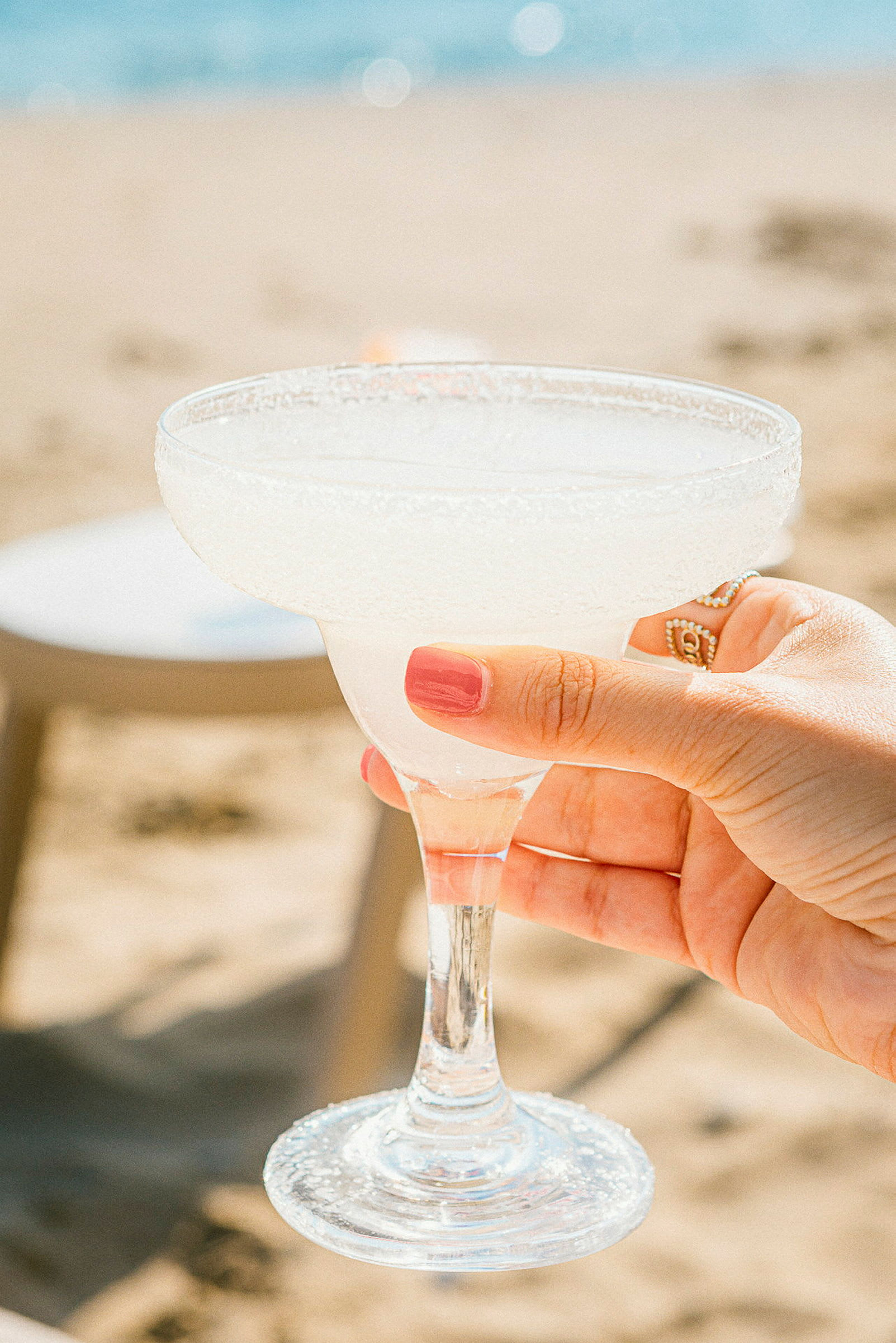 En hand håller ett glas margaritadrink fylld med is bredvid en strand. (AI-genererad text)