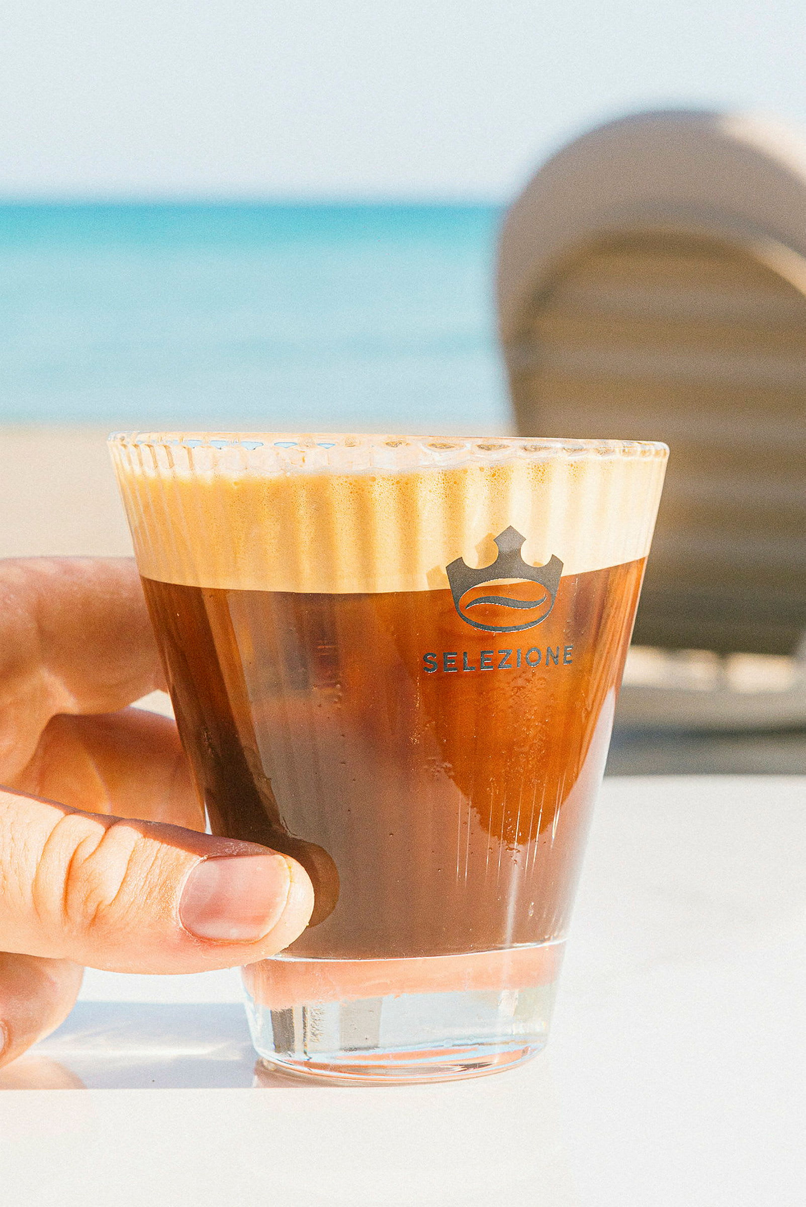 Närbild av en hand som håller ett glas med kaffe, stranden syns i bakgrunden. (AI-genererad text)