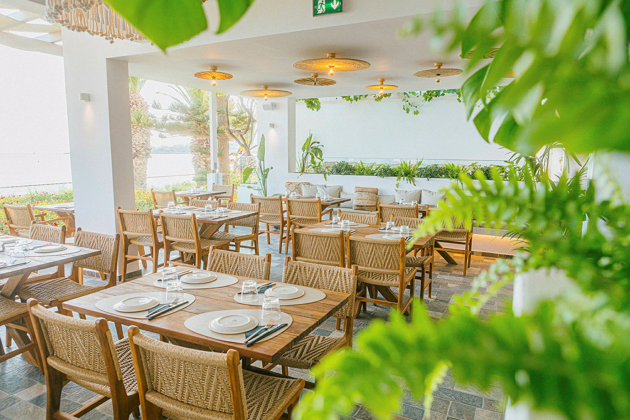En restaurang med bord och stolar i trä, omgiven av grönska och med utsikt över havet. (AI-genererad text)