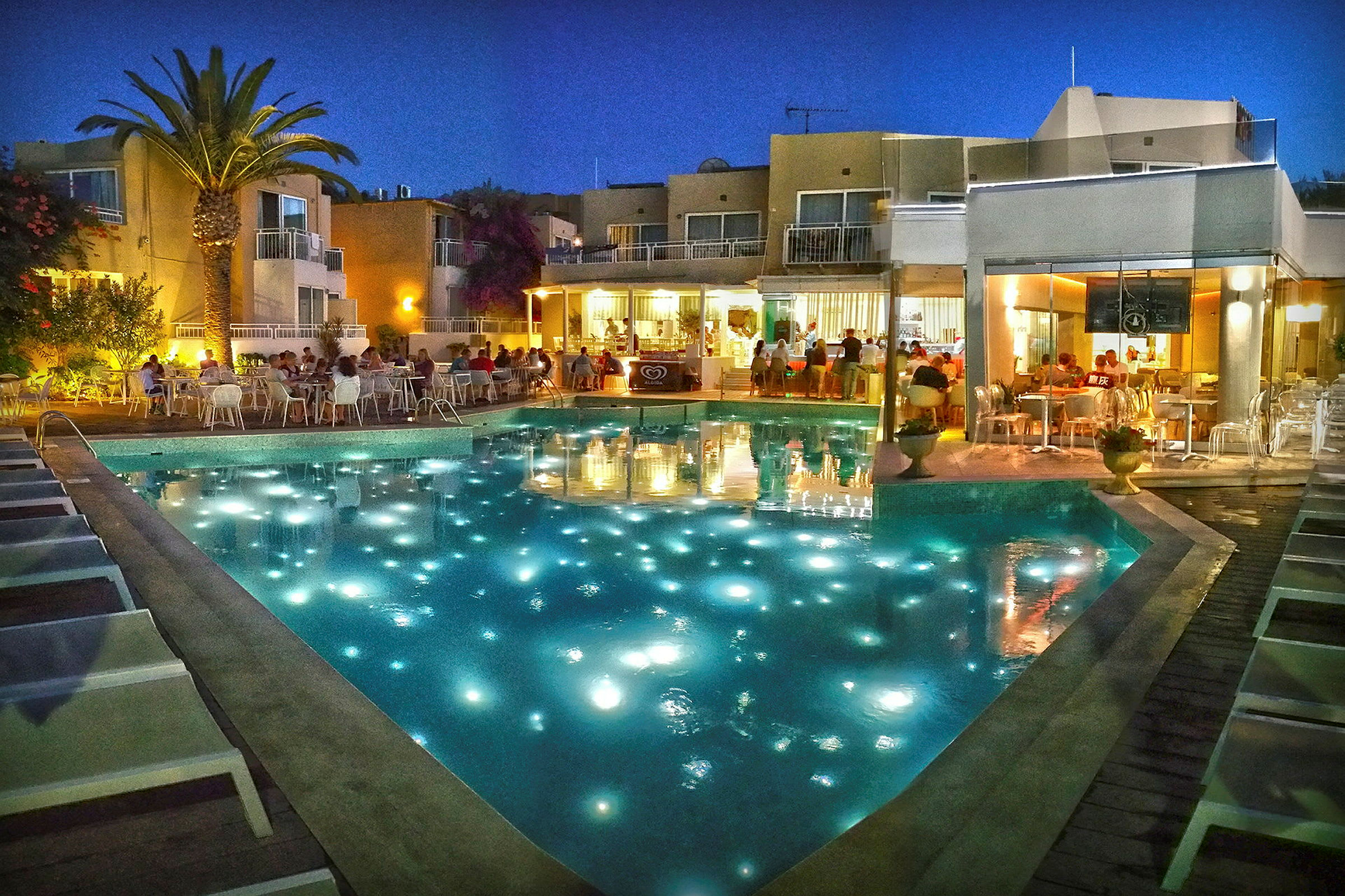 Exteriör av hotellområde med upplyst pool och människor vid restaurang på kvällen. (AI-genererad text)