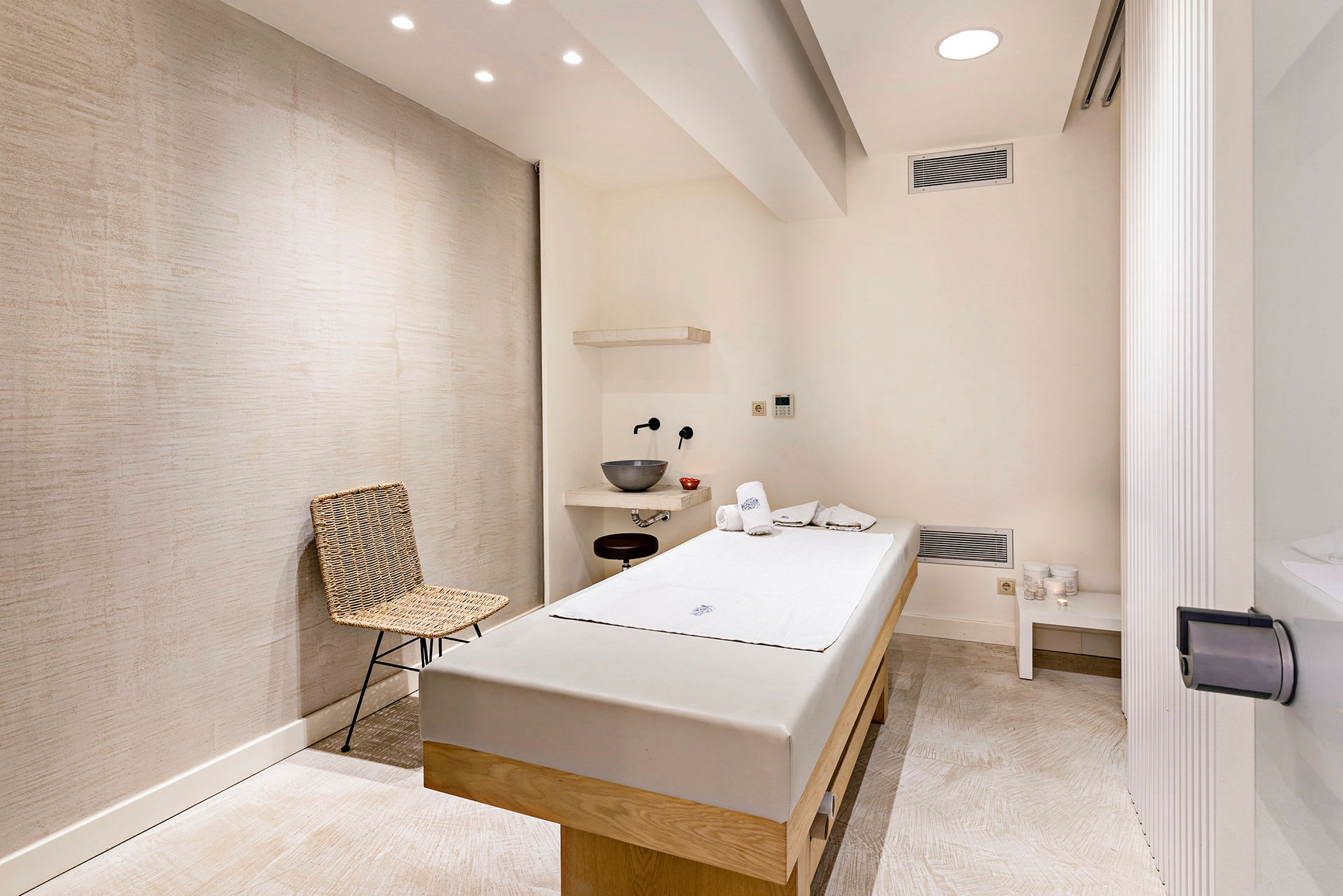 Ett rum för massage med en massagebänk, handdukar och en tvättställ. Dekorativ stol och hyllor på väggen. (AI-genererad text)