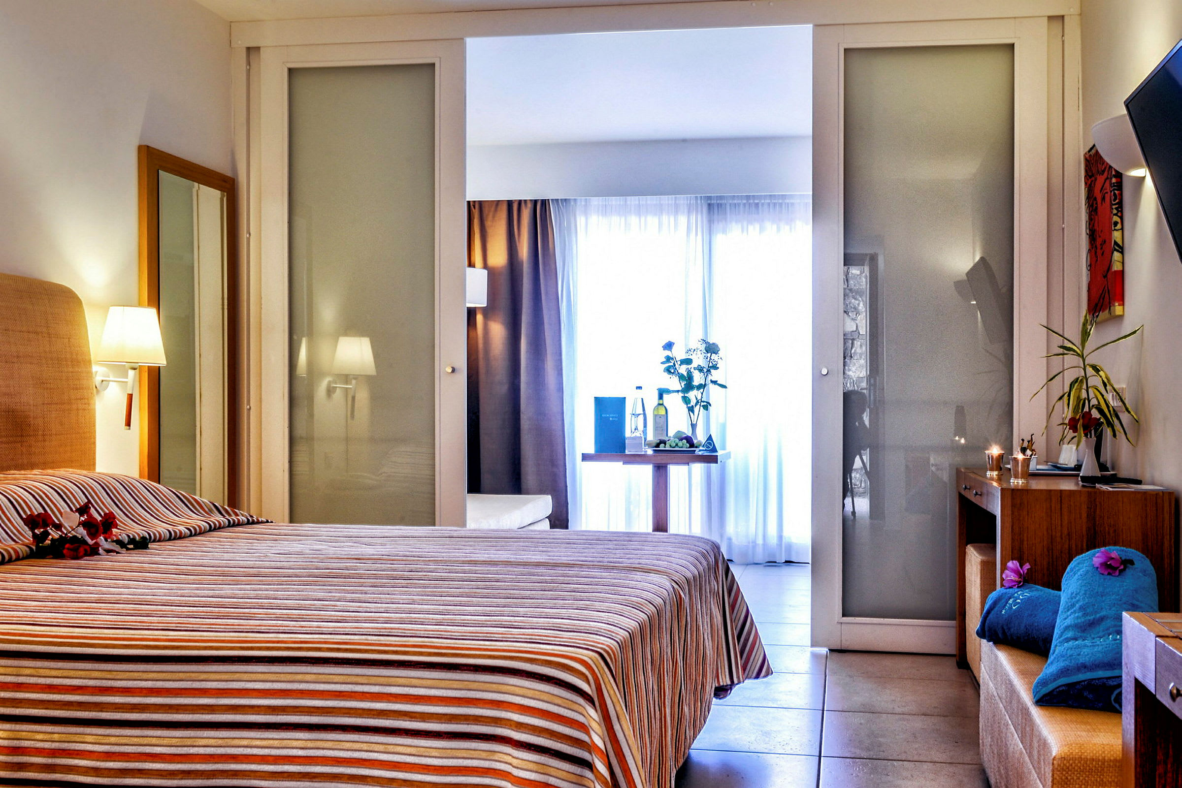 Ett hotellrum med en säng täckt av en randig överkast. I rummet finns även ett bord med dekorativa föremål och bakgrund med glasväggar. (AI-genererad text)