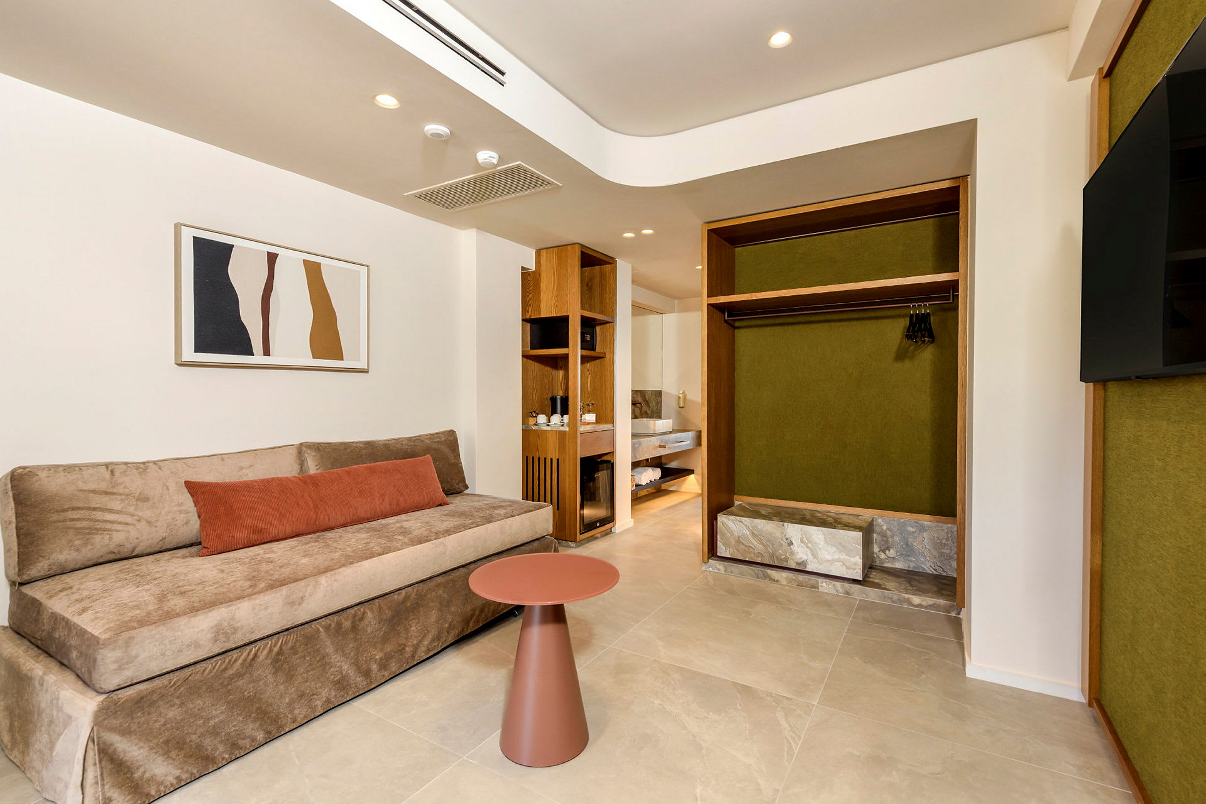 Ett modernt hotellrum med en beige soffa, en orange kudde och ett litet runt bord, sett framför en öppen garderob med gröna paneler. (AI-genererad text)