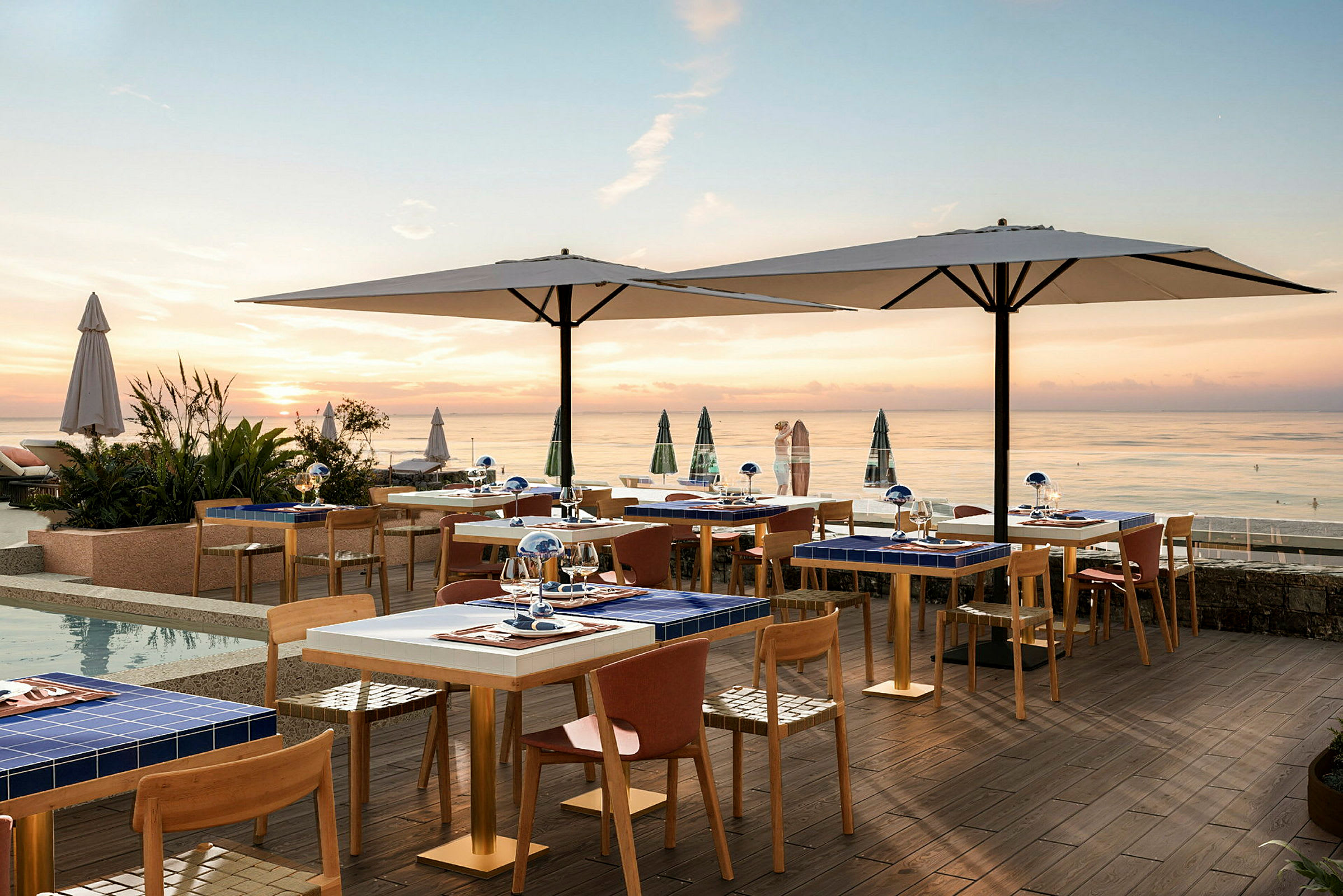 En utomhusrestaurang vid havet under solnedgången med bord och stolar samt stora parasoll. (AI-genererad text)