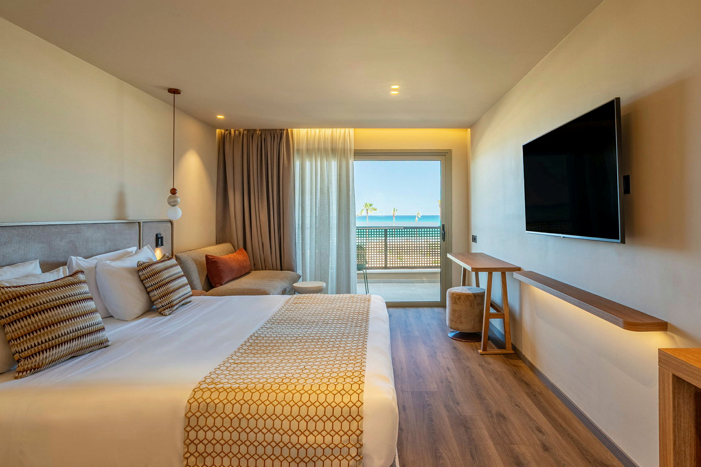 Ett modernt hotellrum med en dubbelsäng, en soffa, en väggmonterad TV och stora fönster med utsikt mot en balkong och havet i bakgrunden. (AI-genererad text)