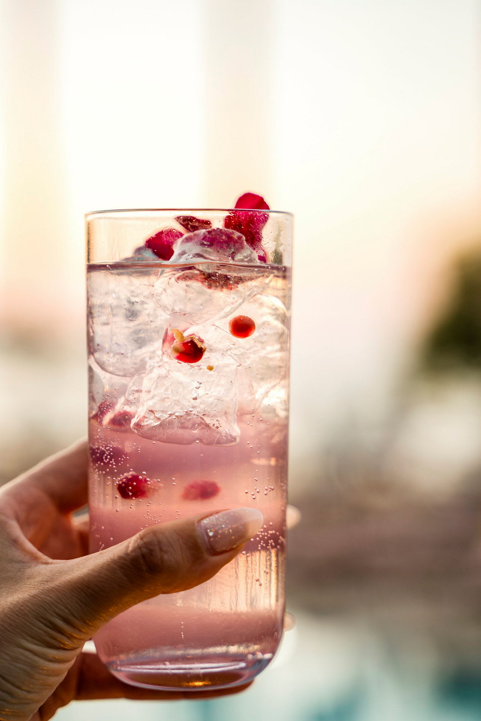Ett glas med rosa dryck, isbitar och granatäpplekärnor hålls av en hand. (AI-genererad text)