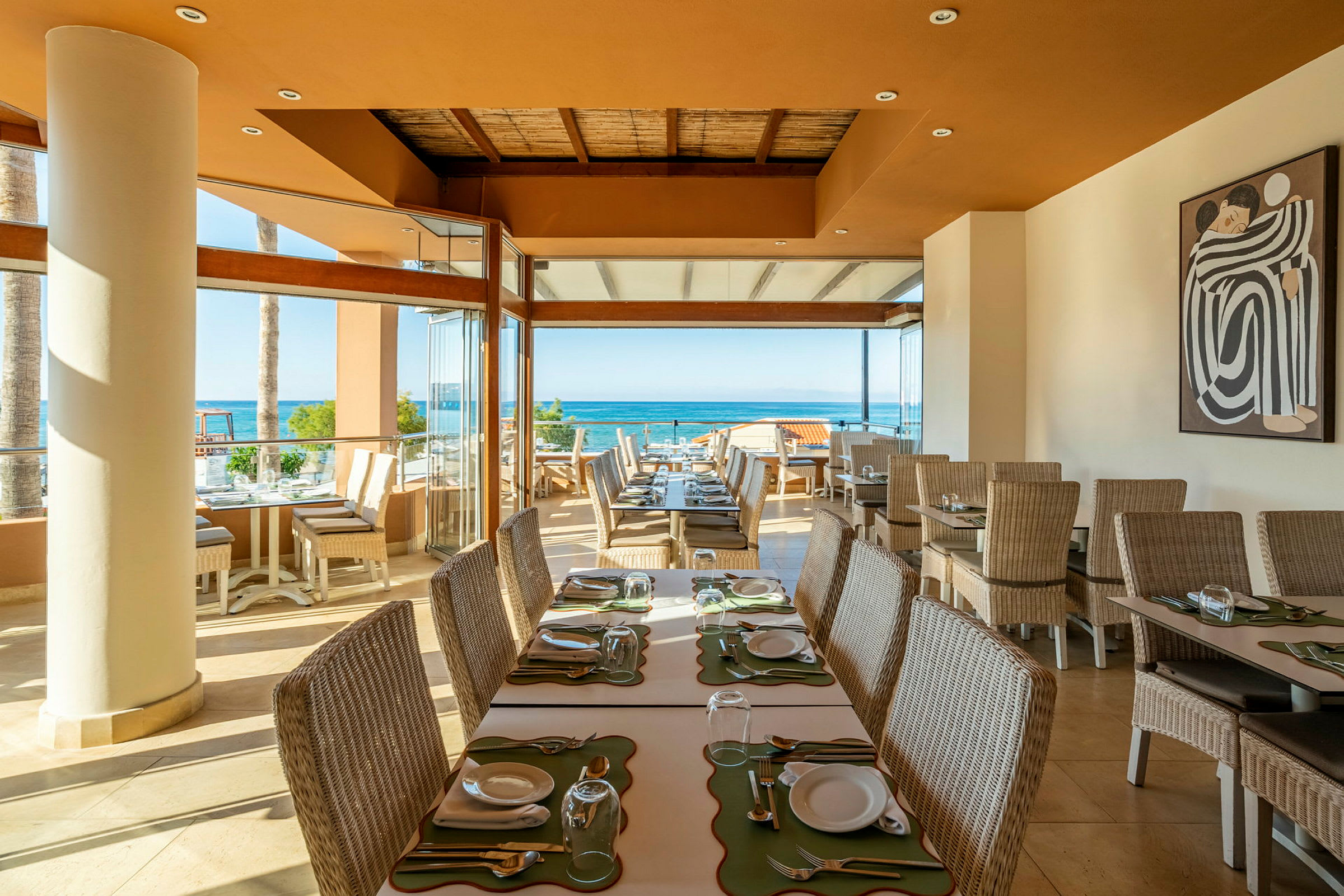 Ett solupplyst restaurangområde med bord och stolar i rotting, utsikt över havet och en målning på väggen. (AI-genererad text)