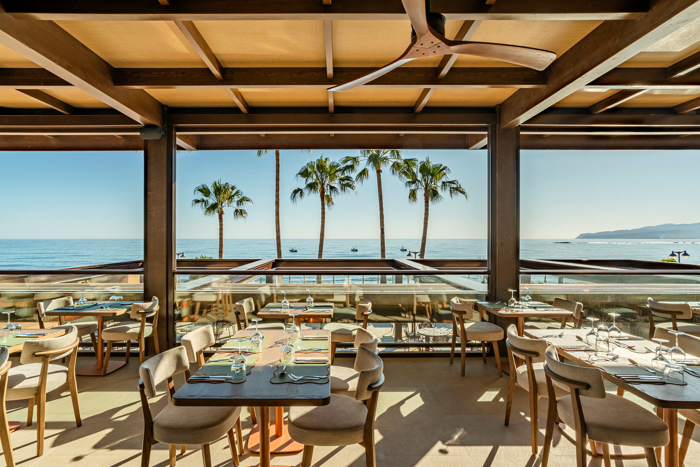 En restaurang med stora fönster och utsikt över havet och palmer. (AI-genererad text)