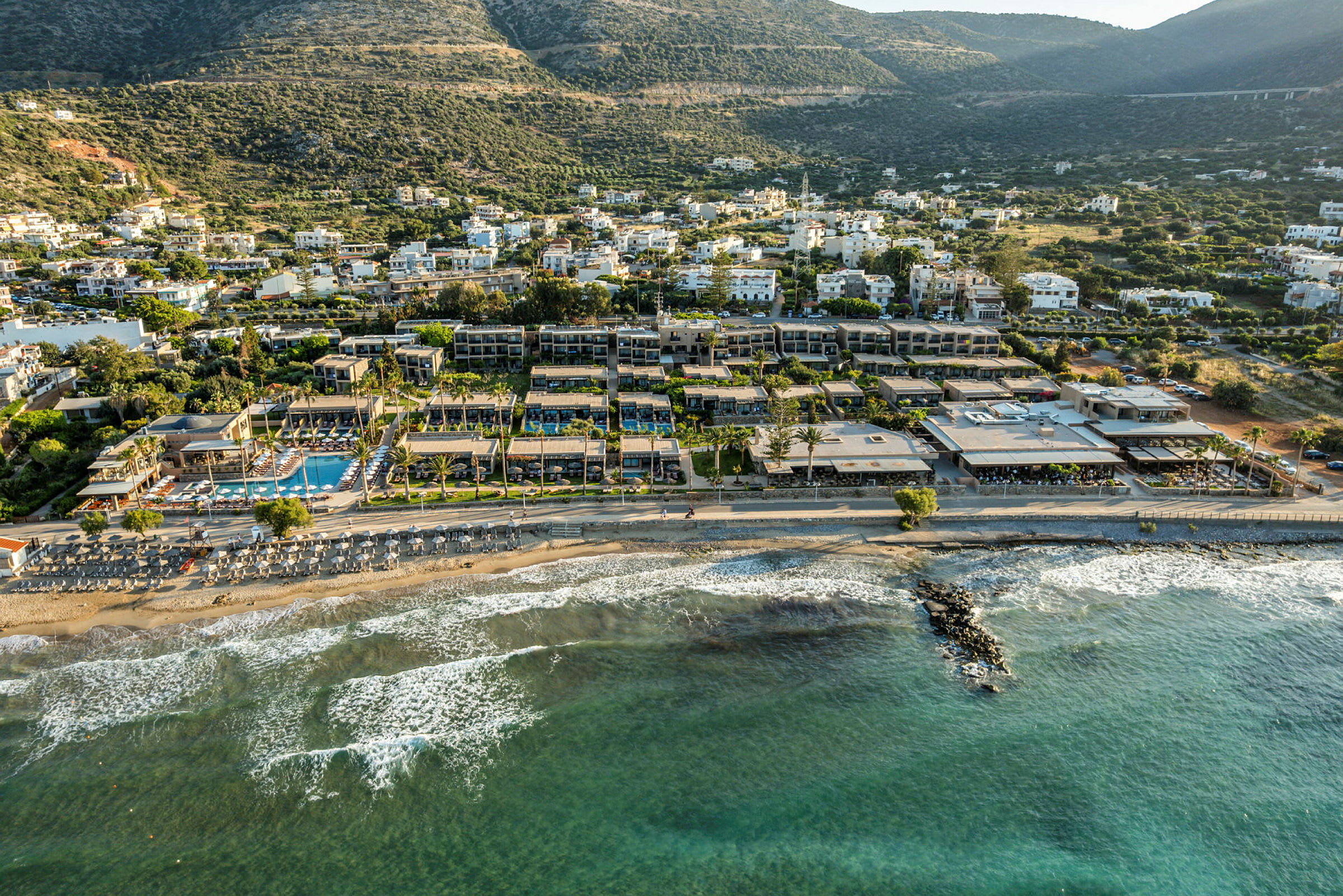 Vy över en strand med solstolar vid vattnet och hotellbyggnader i bakgrunden, med gröna kullar längre bort. (AI-genererad text)
