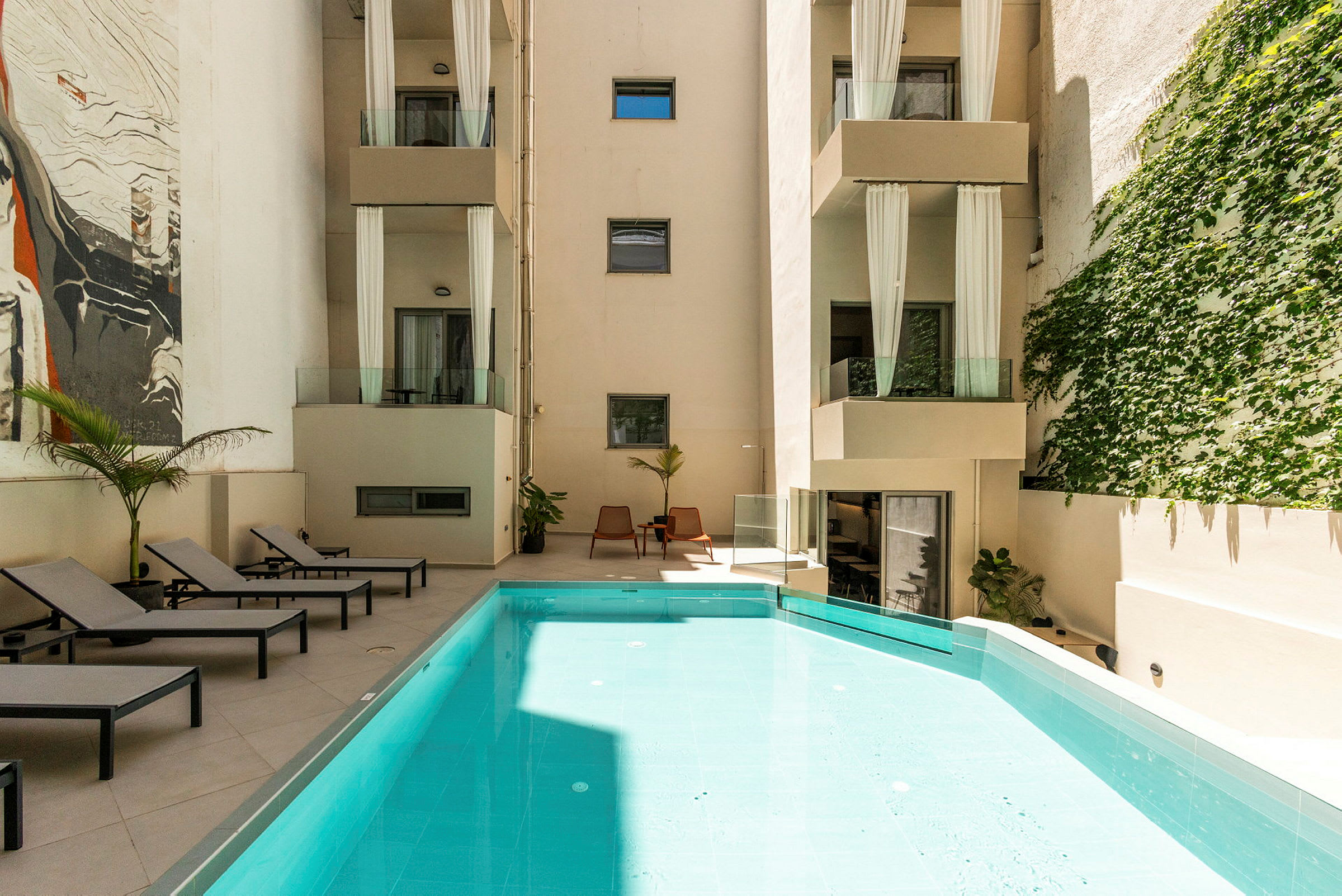 En modern byggnad med flera balkonger och en pool i förgrunden. Solstolar är placerade vid sidan, och växtlighet täcker en del av väggen. (AI-genererad text)