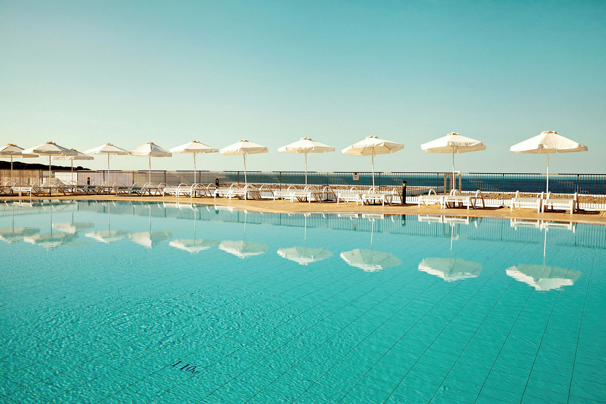 Utomhuspool med klara vatten reflekterande vita parasoller, solstolar och havet i bakgrunden. (AI-genererad text)