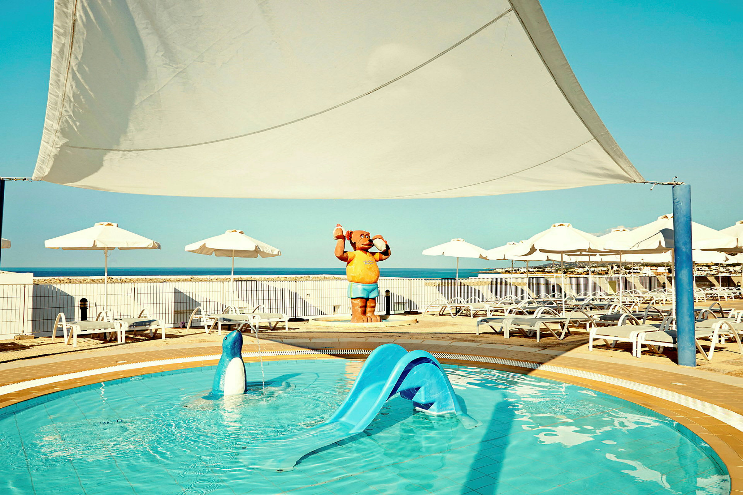 En pool med rutschkana omgiven av solstolar och parasoller nära havet. (AI-genererad text)