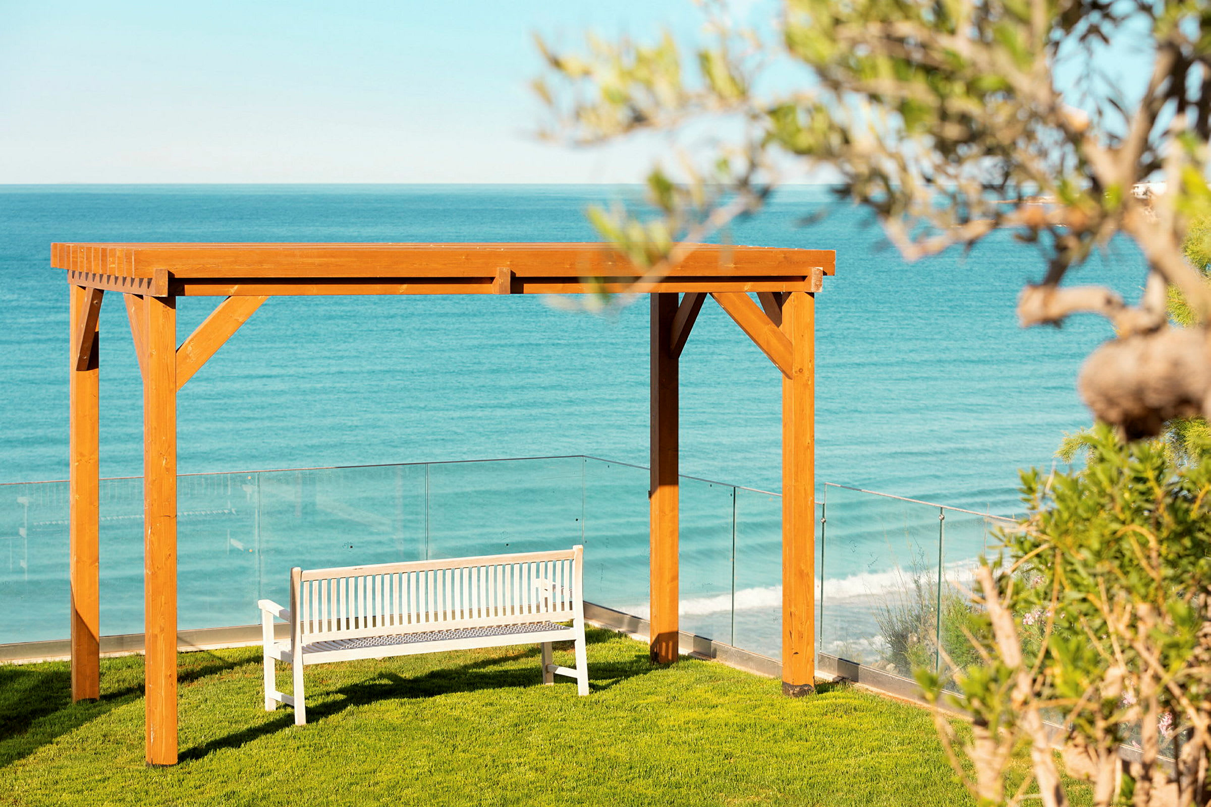 En vit bänk under en träpergola vid en gräsmatta med havet i bakgrunden. (AI-genererad text)