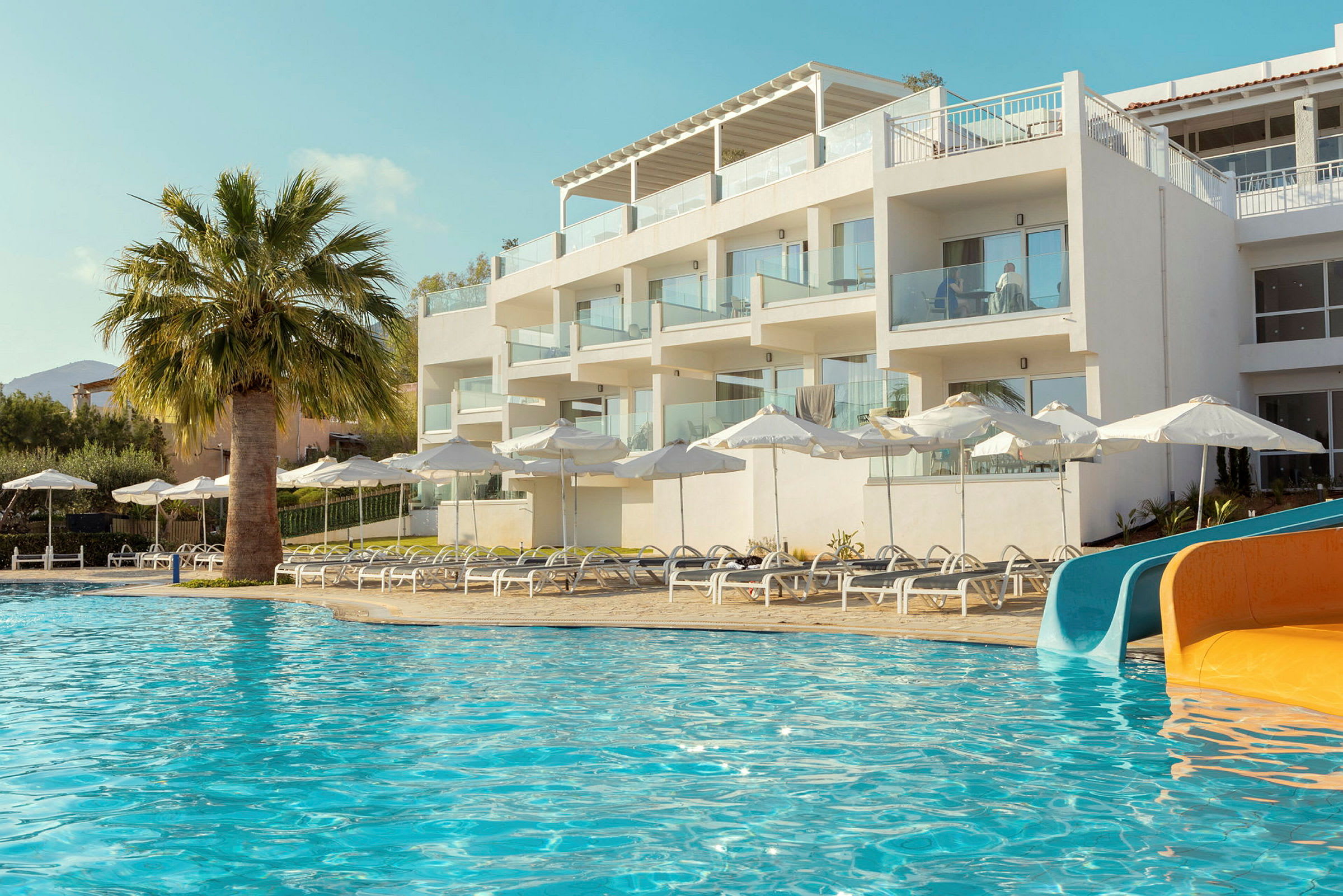 Ett hotell med vita byggnader vid en pool med solstolar, parasoller och vattenrutschkanor. (AI-genererad text)
