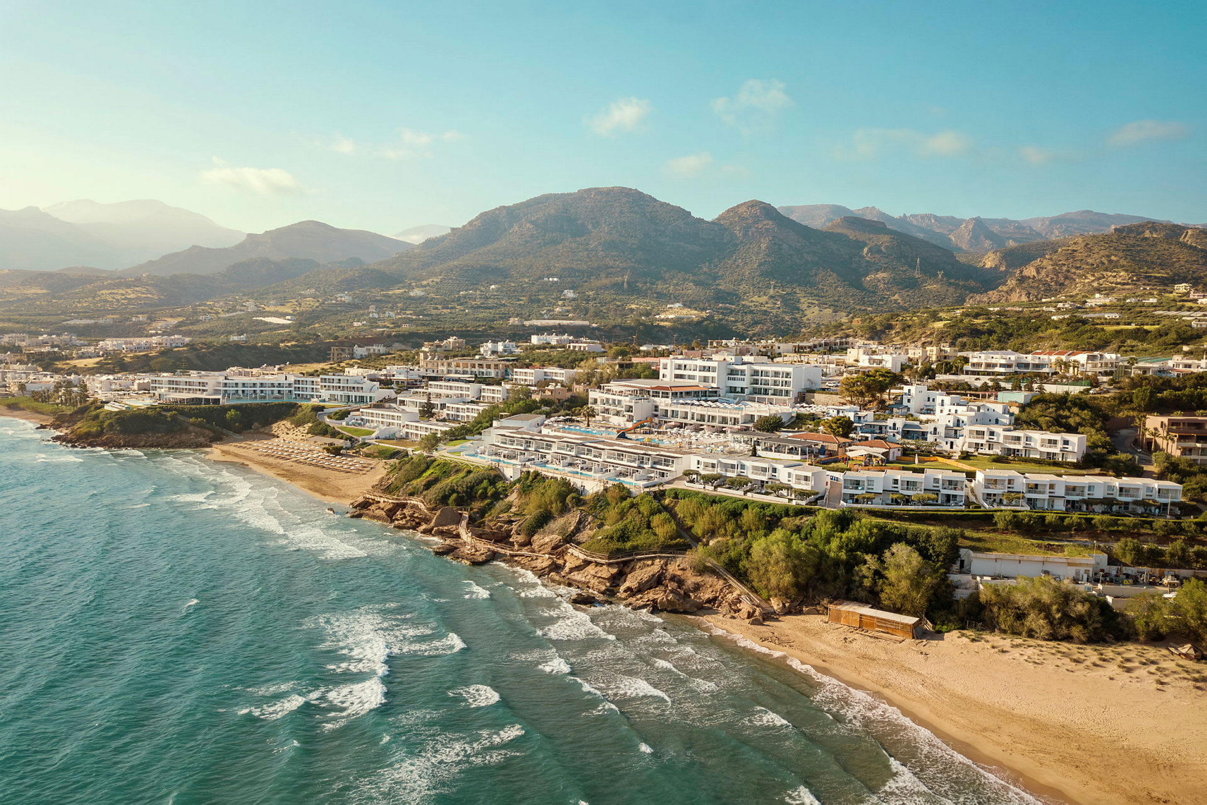 Utsikt över en kustlinje med turkost hav, sandstrand och hotellbyggnader omgivna av grönska och berg i bakgrunden. (AI-genererad text)