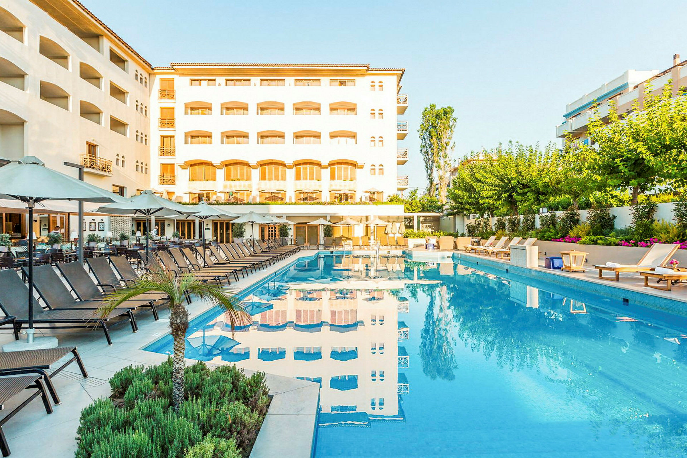 En utomhuspool med omgivande solstolar och parasoller framför ett hotell. (AI-genererad text)