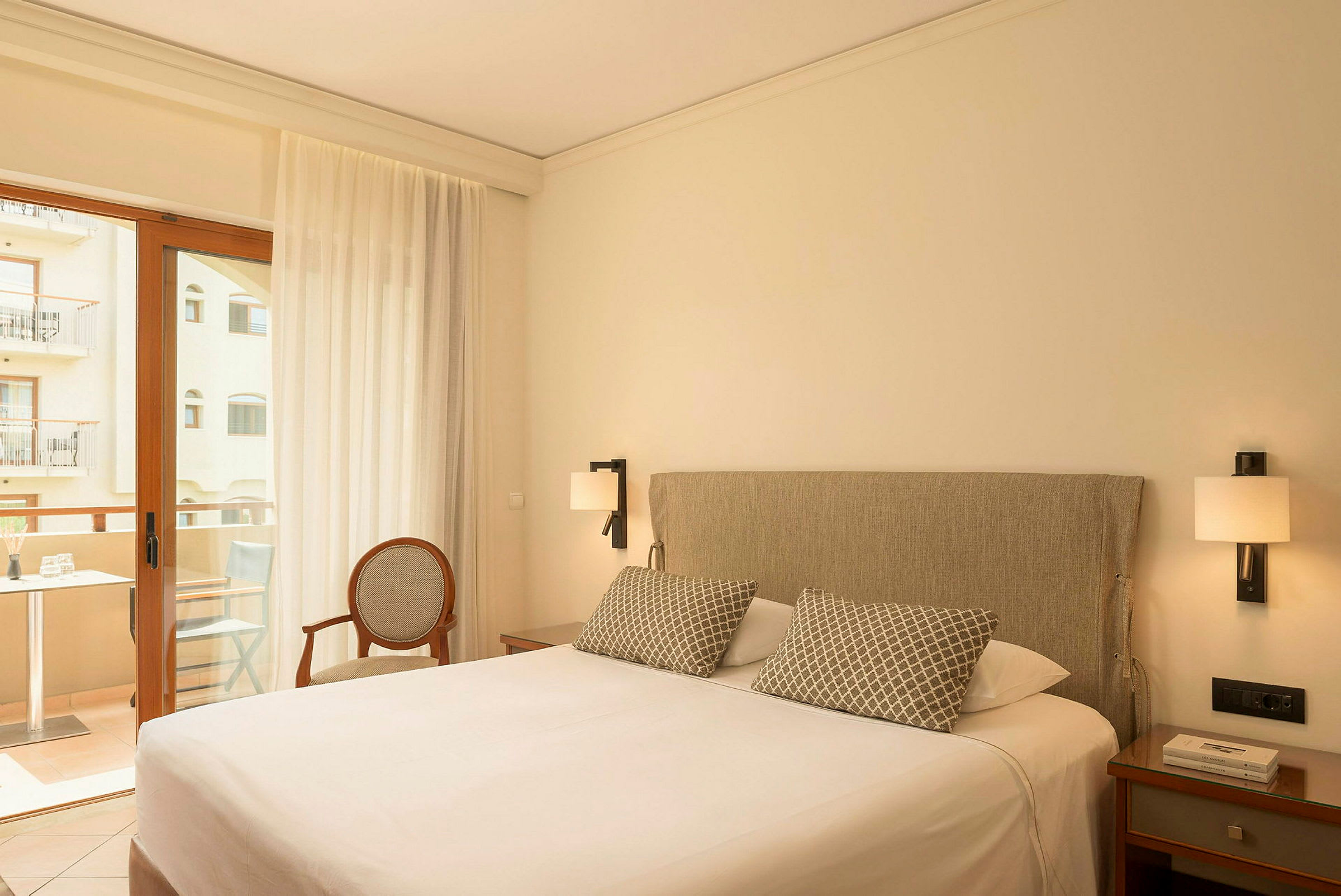 Hotellrum med en dubbelsäng, nattduksbord, en stol och en altandörr med utsikt mot en balkong. (AI-genererad text)