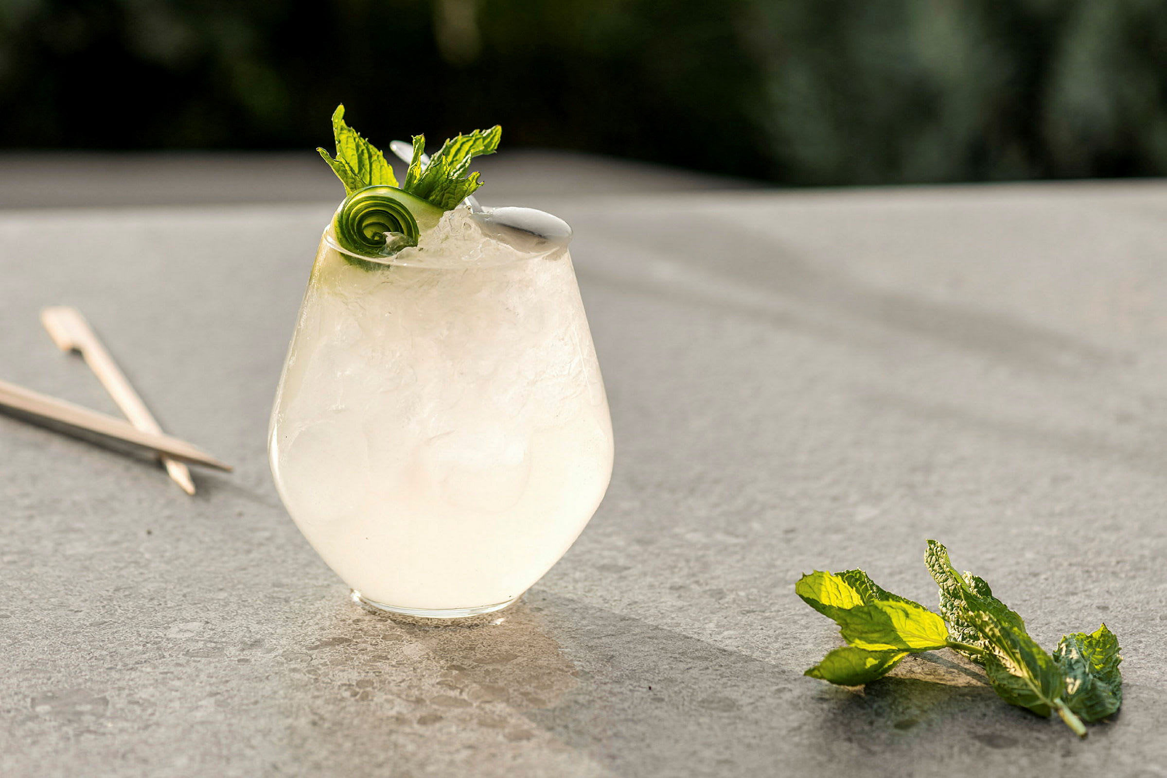 Ett glas med en kall drink, garnerat med mynta och gurkspiral, står på en bordsyta. (AI-genererad text)