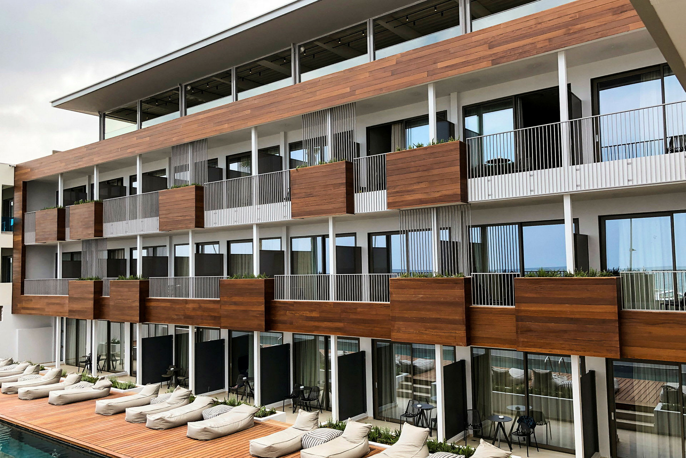 Hotellbyggnad med träpanel och balkonger, med solsängar vid poolen. (AI-genererad text)