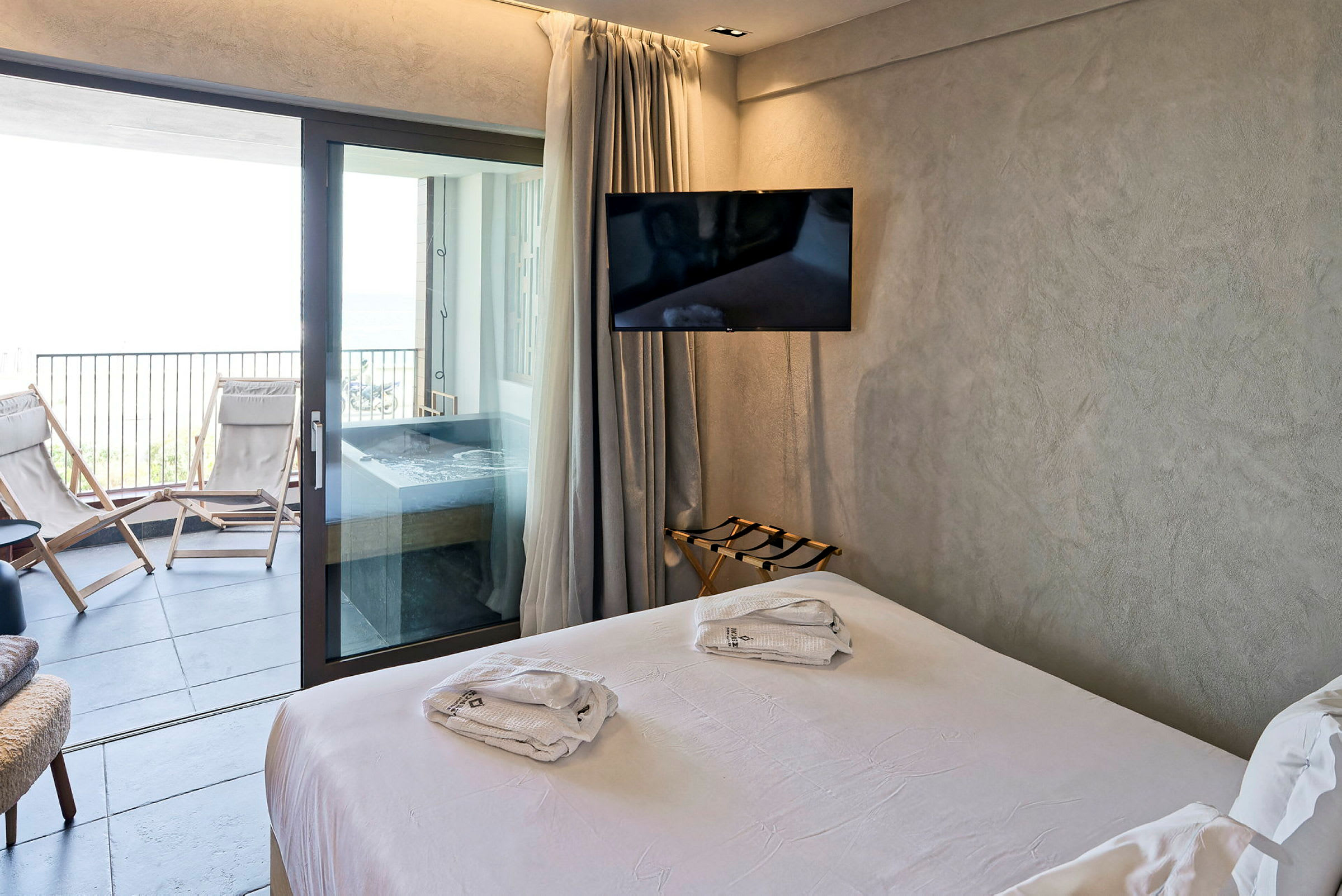Modernt hotellrum med vit dubbelsäng som leder ut till en balkong med vilstolar och jacuzzi. (AI-genererad text)