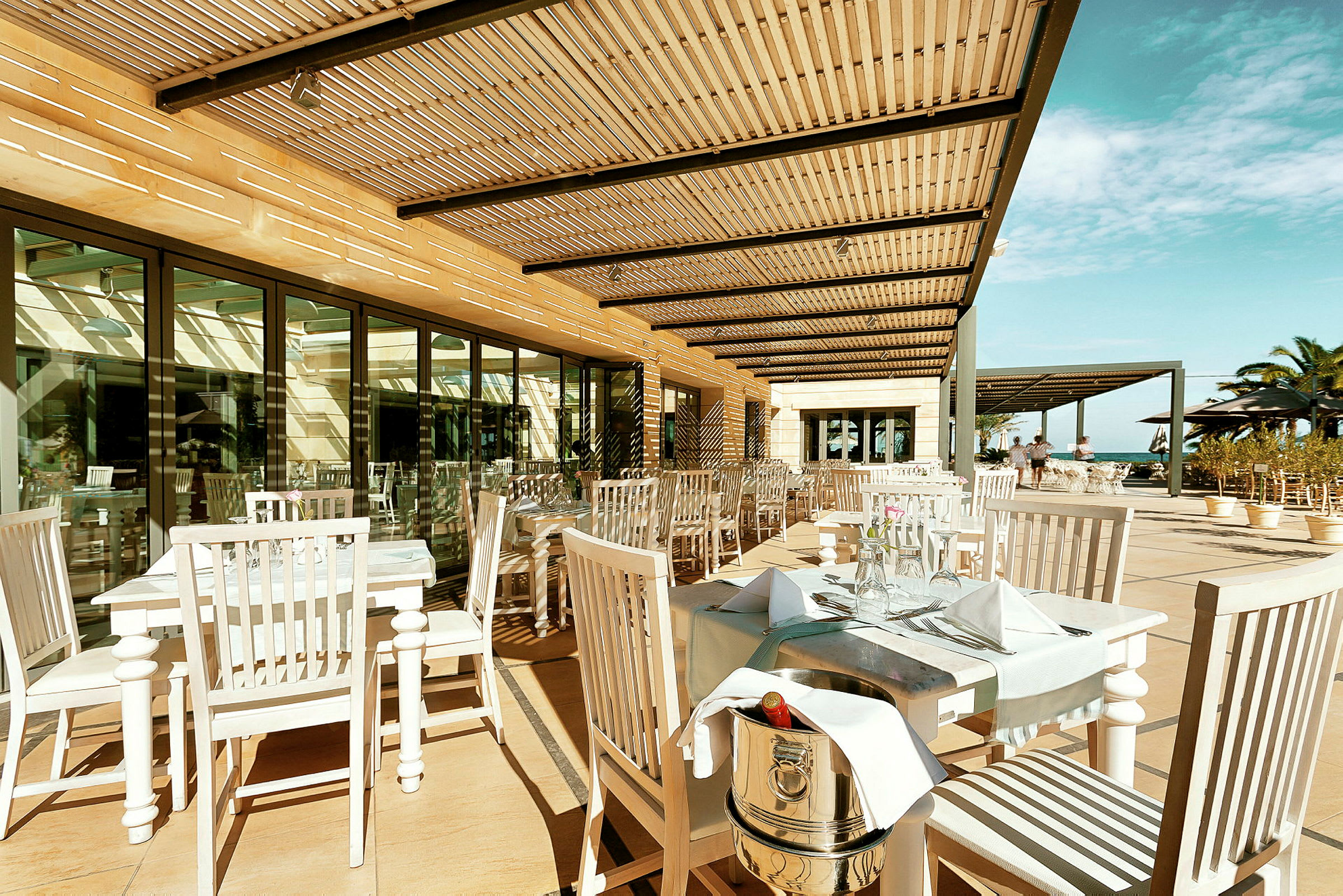 Utomhusrestaurang med vita bord och stolar samt pergola som skuggar. (AI-genererad text)