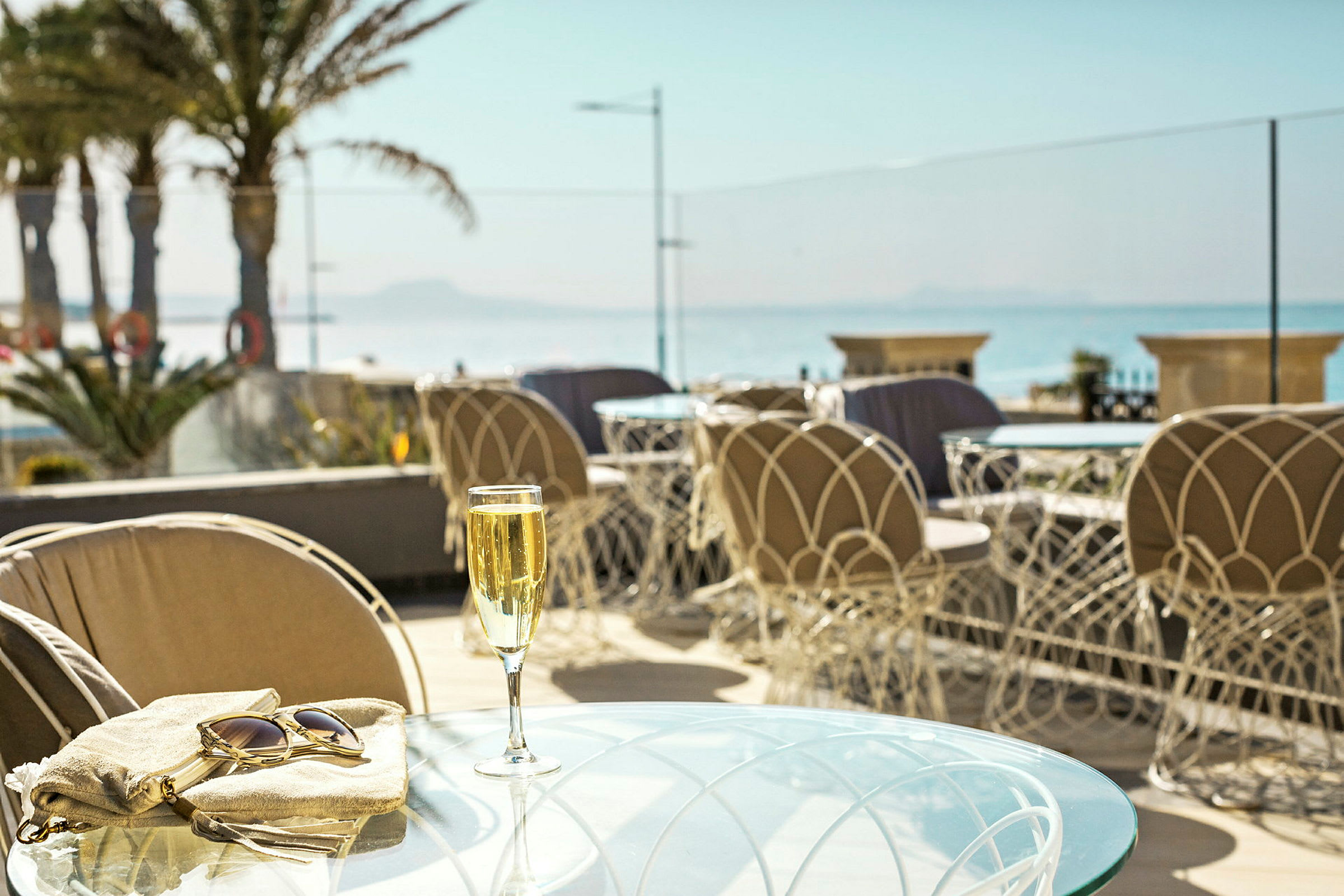 Ett glas champagne på ett bord, med en handväska och solglasögon bredvid, framför en utsikt över havet och palmer. (AI-genererad text)