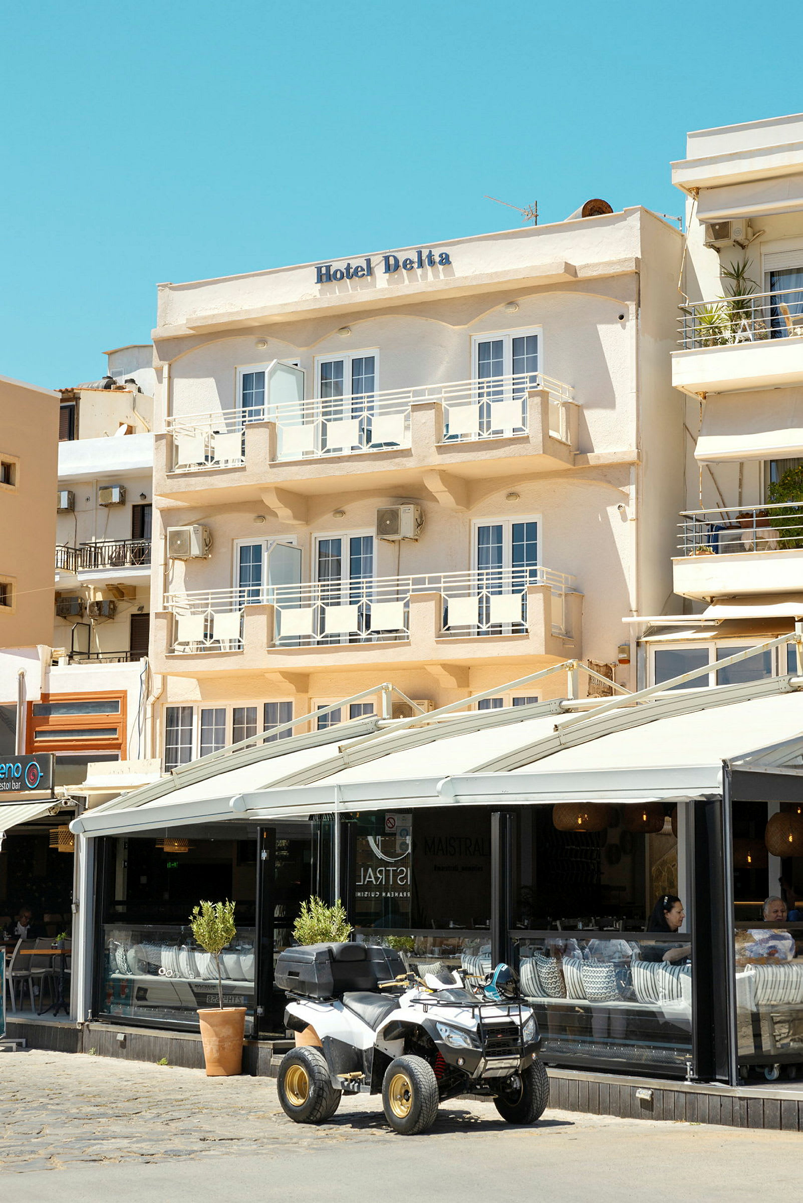 Fasad av ett hotell med namnet "Hotel Delta" med balkonger och ett café på bottenvåningen, där en fyrhjuling står parkerad utanför. (AI-genererad text)