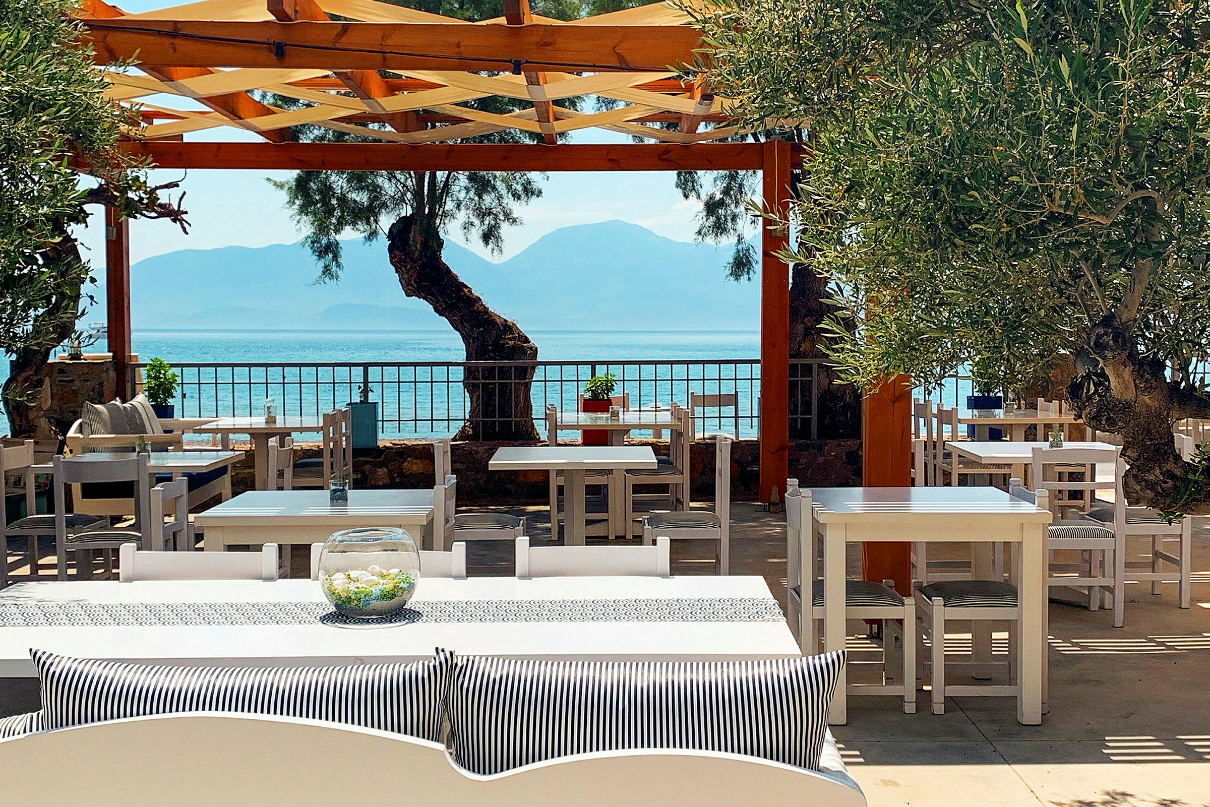 Bild av en utomhusrestaurang med vita bord och stolar, havet och berg synliga i bakgrunden. (AI-genererad text)