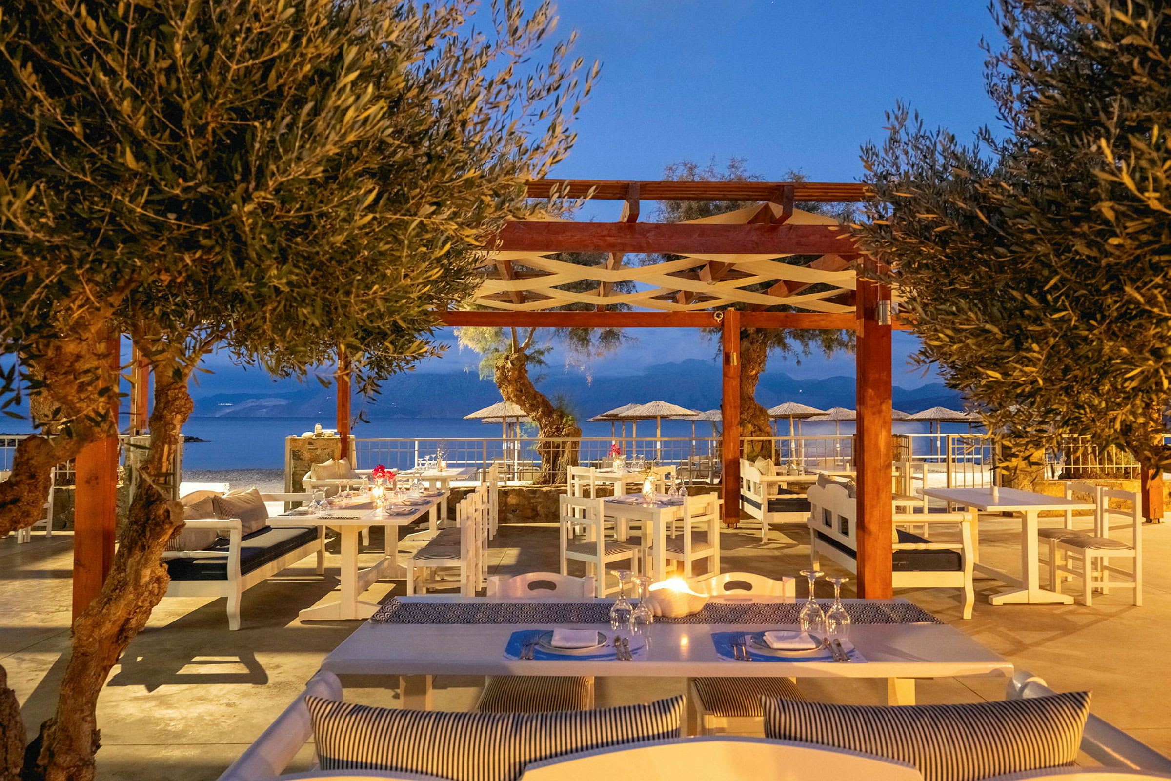 En utomhusrestaurang med sittplatser under en träpergola och utsikt över havet. (AI-genererad text)