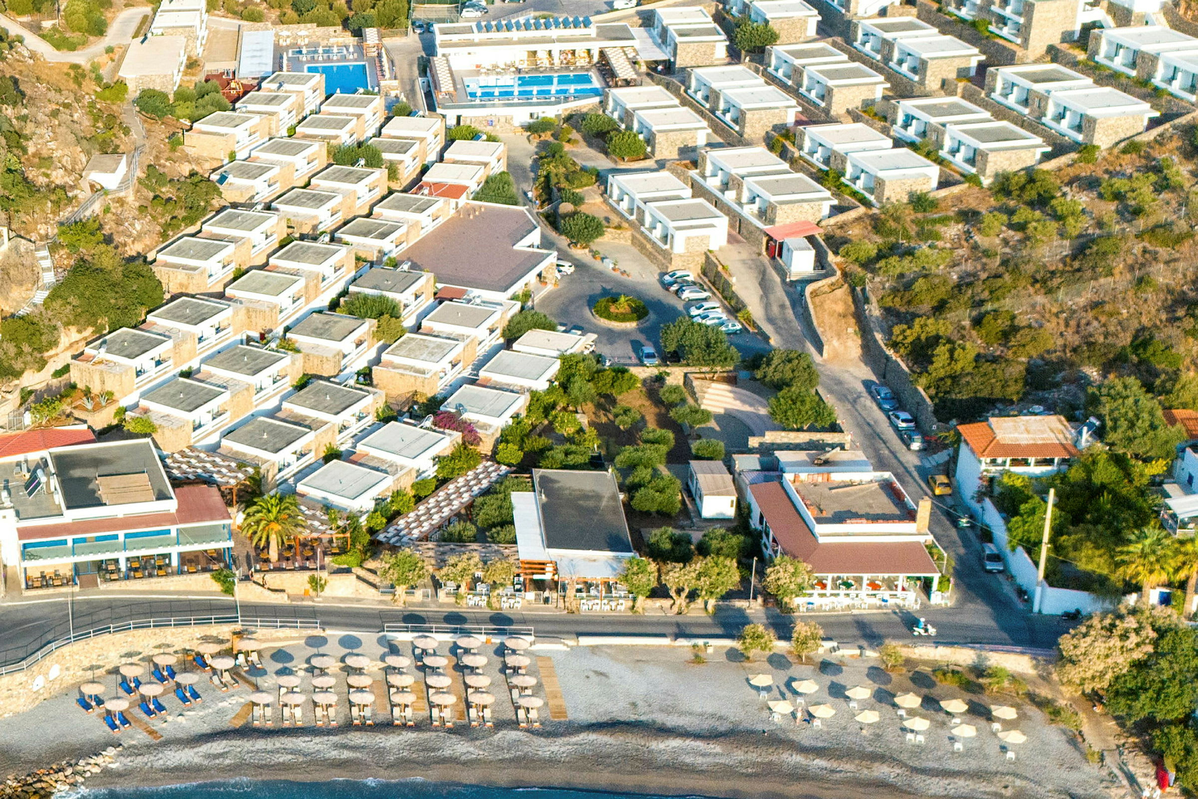 Flygfoto av en semesteranläggning med byggnader, pooler och en strand med parasoller. (AI-genererad text)