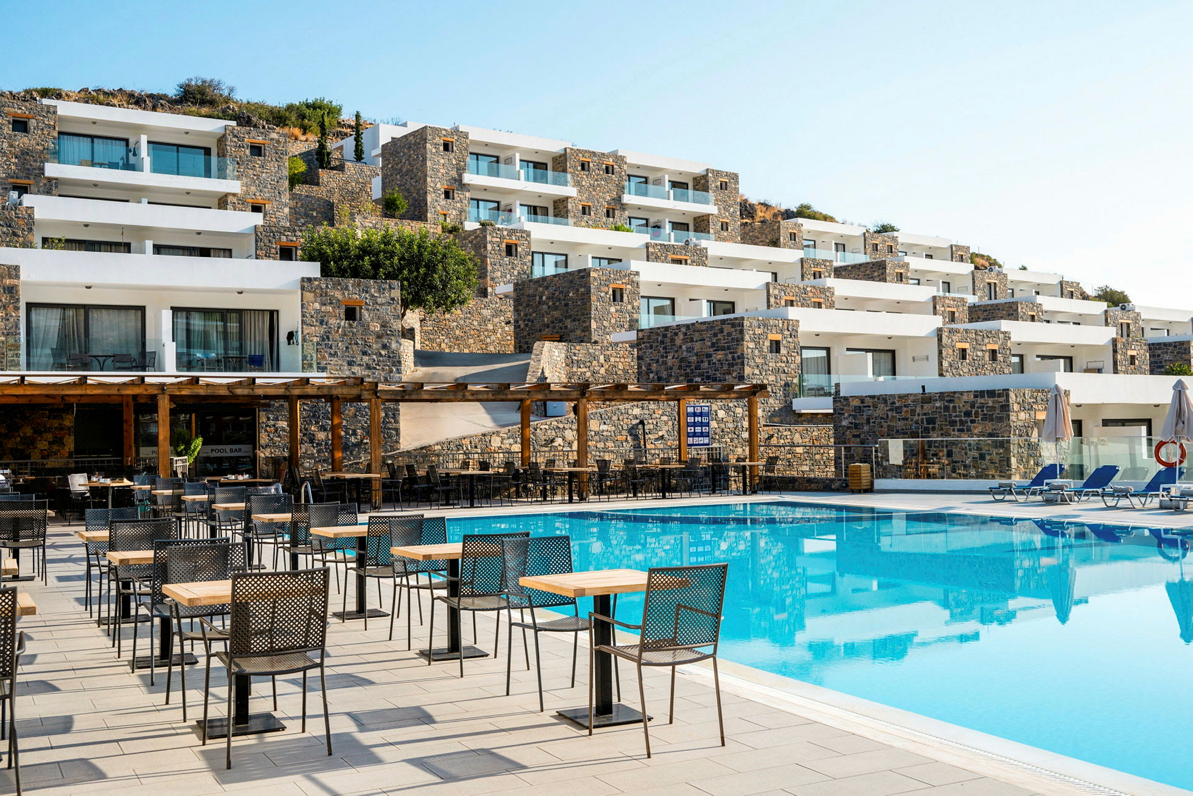 Bild av hotellets utomhusområde med pool, bord och stolar samt stenbyggnader i bakgrunden. (AI-genererad text)