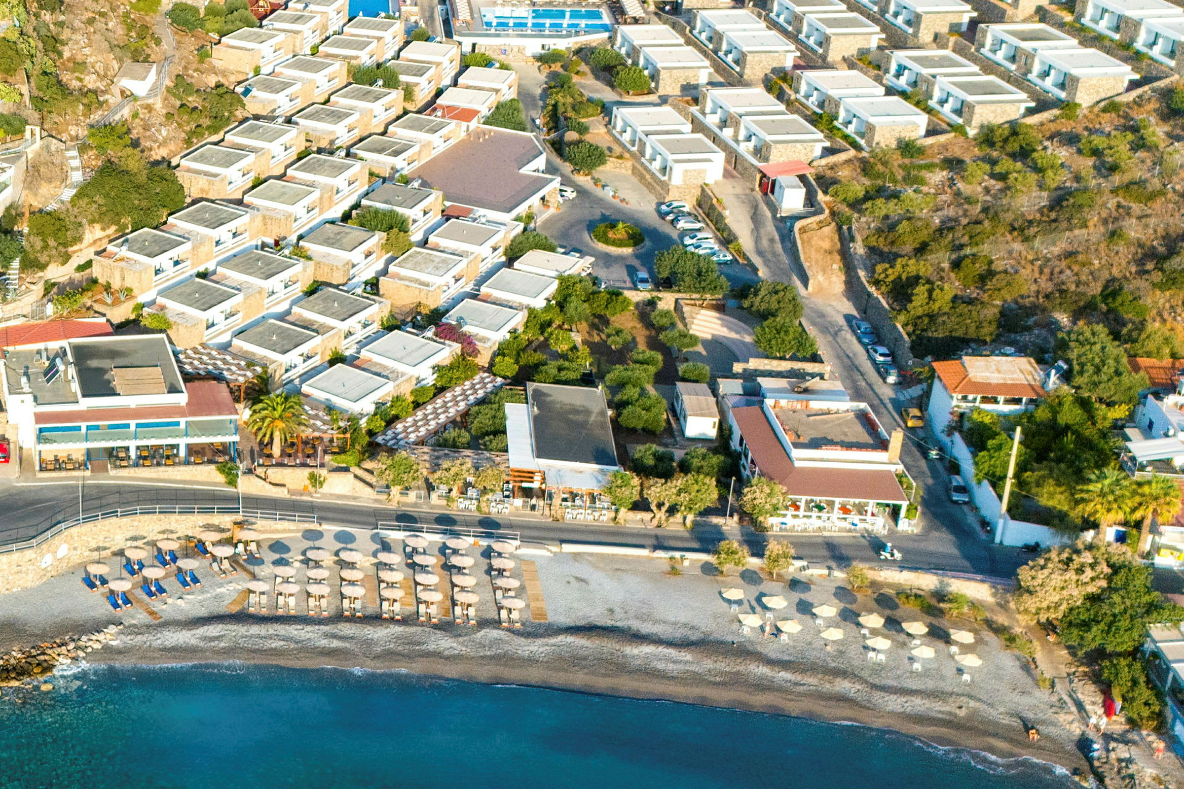 Flygfoto av hotellområde med pool, bilparkering och närliggande strand med parasoller. (AI-genererad text)