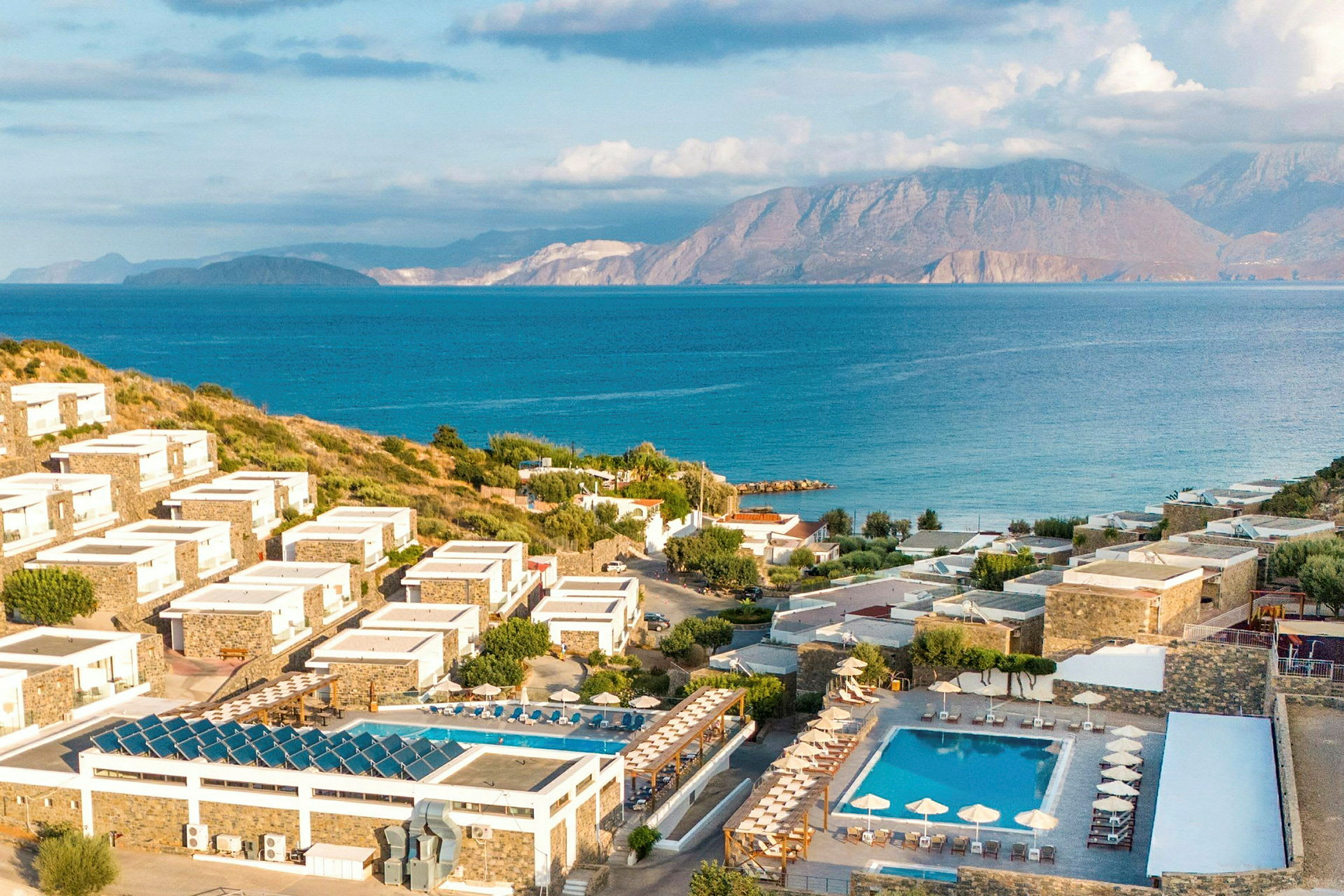Bild av en kustnära resort med hotellbyggnader på sluttande mark omgivna av gröna områden och pooler, med hav och berg i bakgrunden. (AI-genererad text)